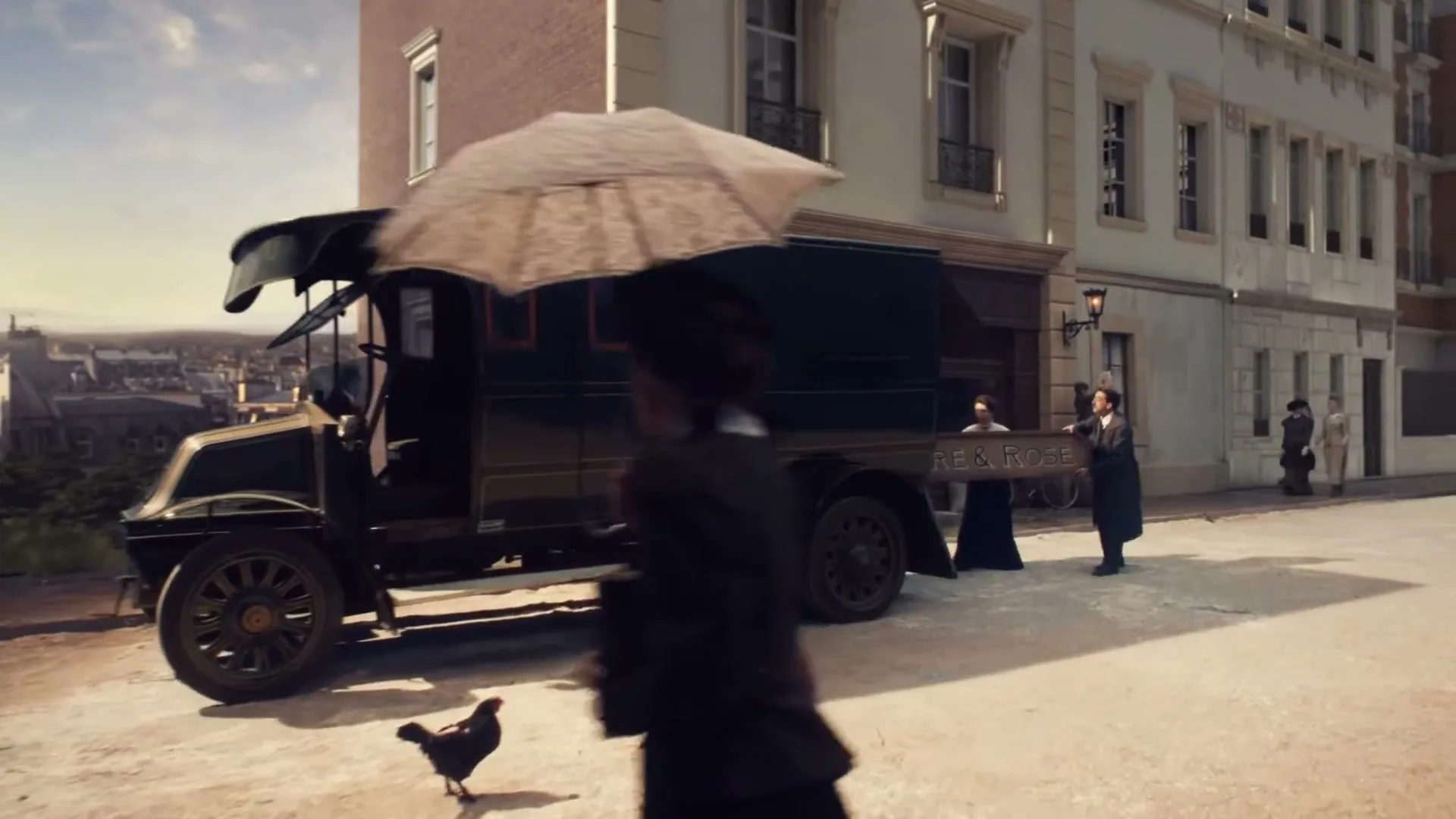 A vintage black delivery truck is parked on a cobblestone street in a European town, as people walk by and chickens peck at the ground.