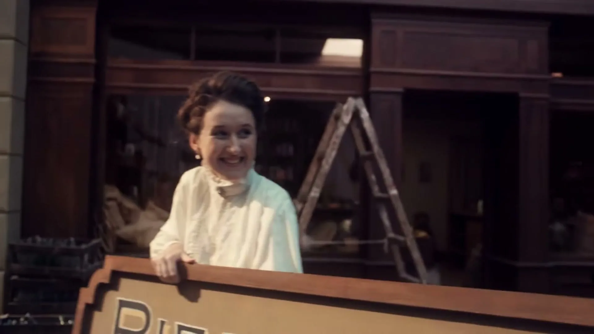 A man with a mustache and glasses struggles to carry a large sign out of a store, while a woman behind him smiles in a bustling city street.