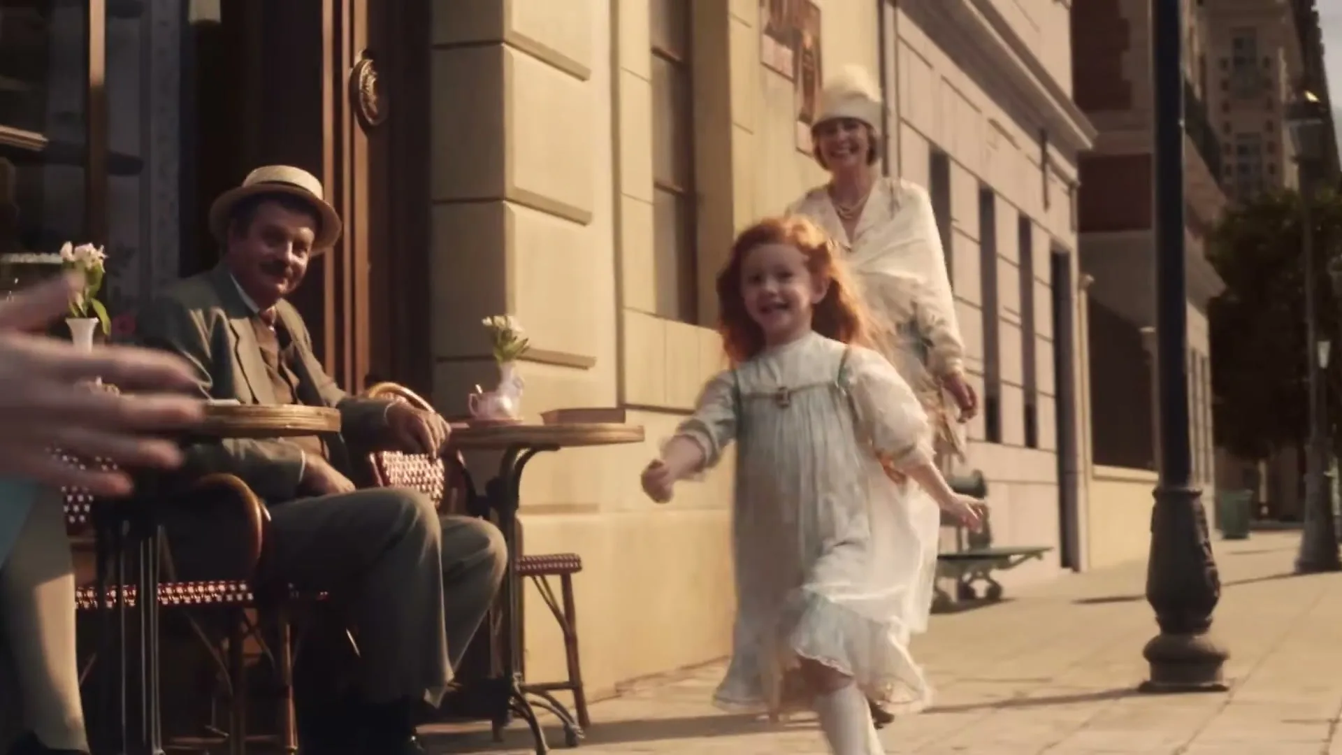 A young girl with red hair runs joyfully on a sidewalk in a European city, while an older woman watches her and a man sits at a cafe table.
