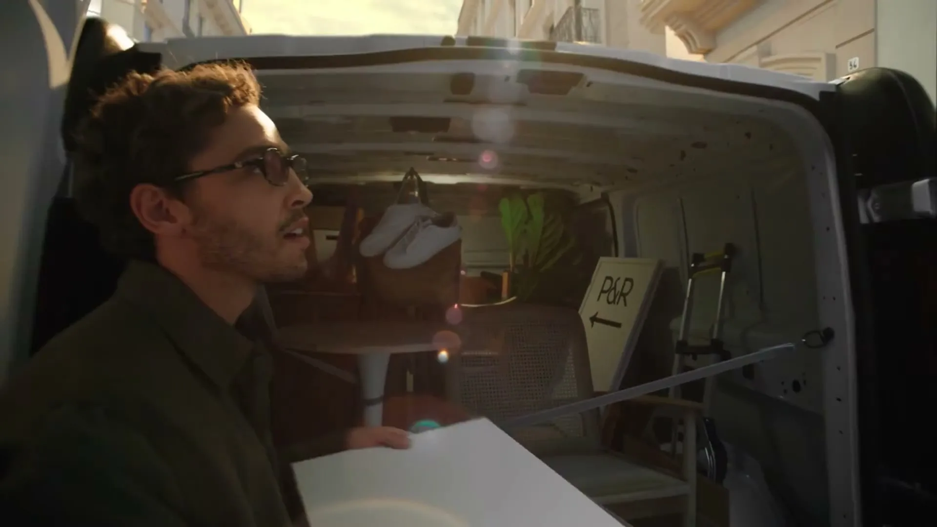 A young man with glasses and a green shirt looks up and to the right, holding a white board, standing in front of a white van with its back doors open, revealing furniture and a "P&R" sign inside, on a sunny street.