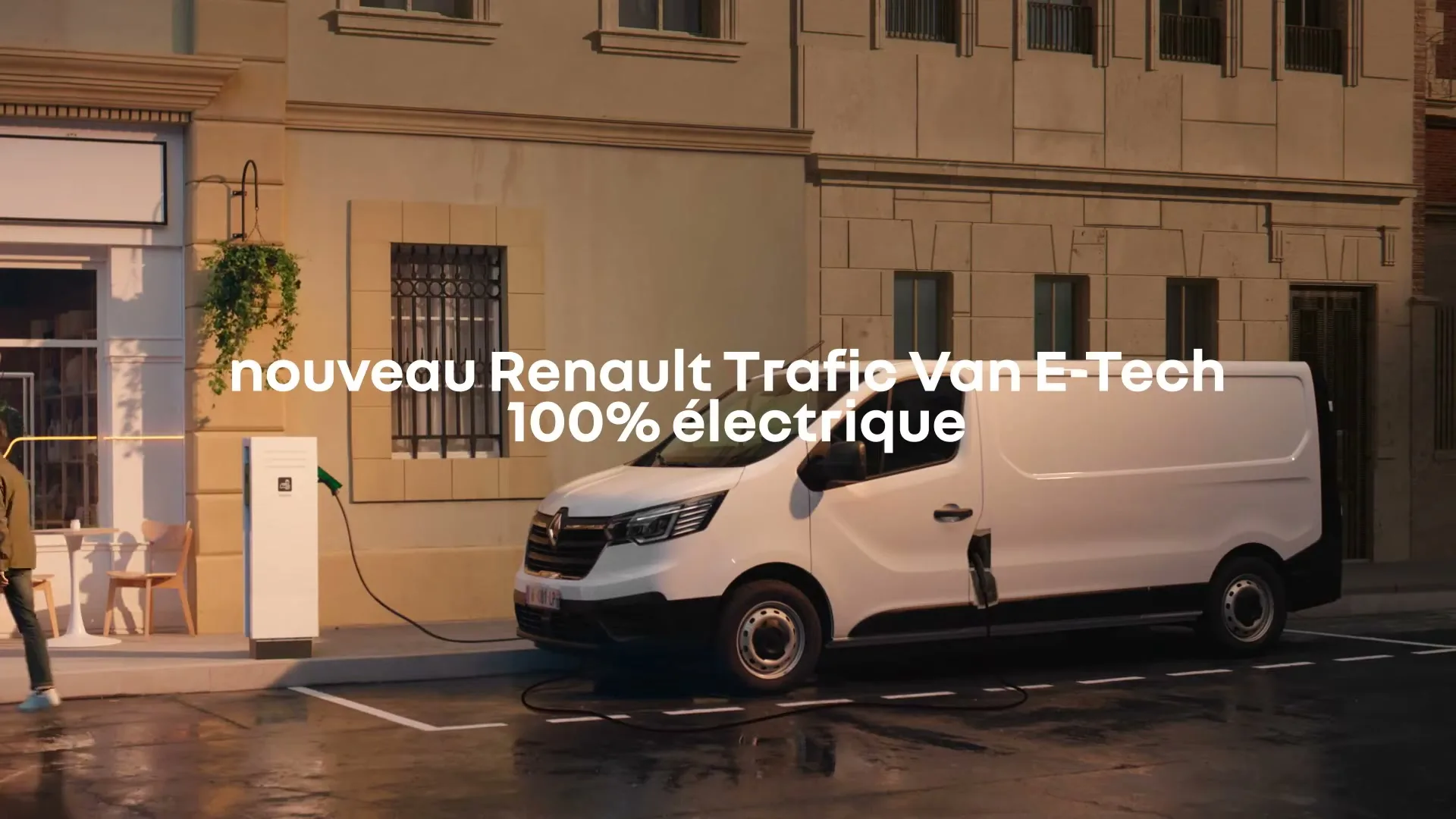 A man in a jacket and sunglasses walks past a white electric Renault Trafic van charging on a city street at dusk.