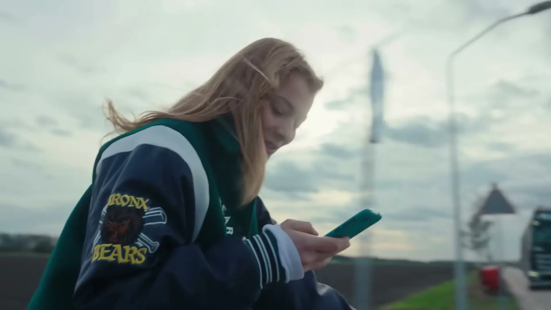 A young woman with blonde hair, wearing a varsity jacket, smiles while looking at her phone on the side of a road, then a truck passes by.
