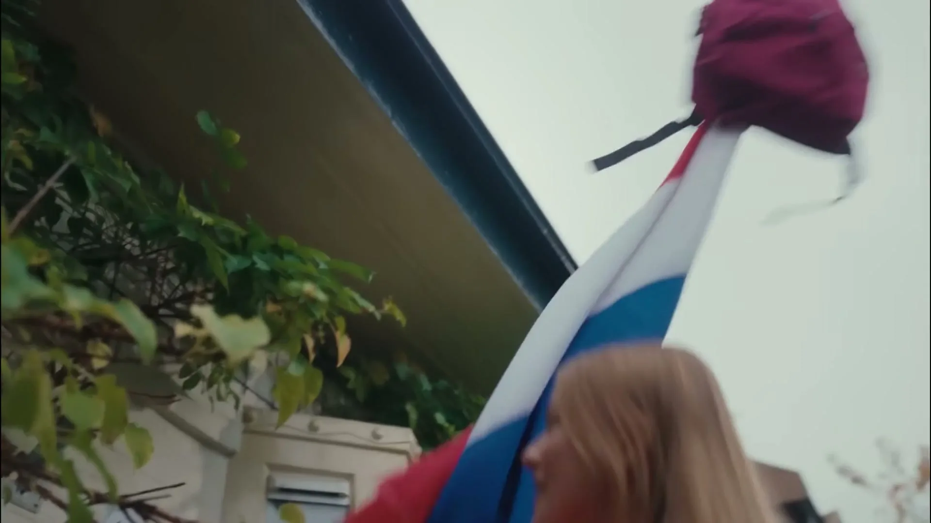 A young woman with long blonde hair, wearing a pink top, struggles to put a red backpack on top of a flagpole with a Dutch flag, outside a house.
