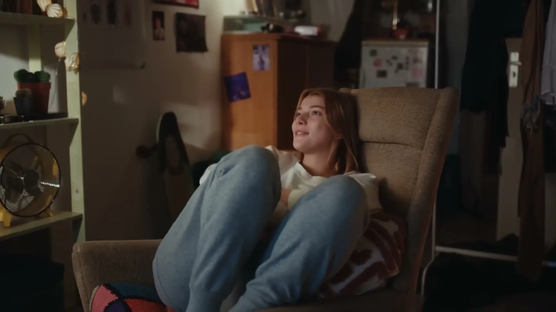 A young woman with light brown hair, wearing a white sweatshirt and grey sweatpants, sits in a brown armchair, looking up and to the right, in a dimly lit room.
