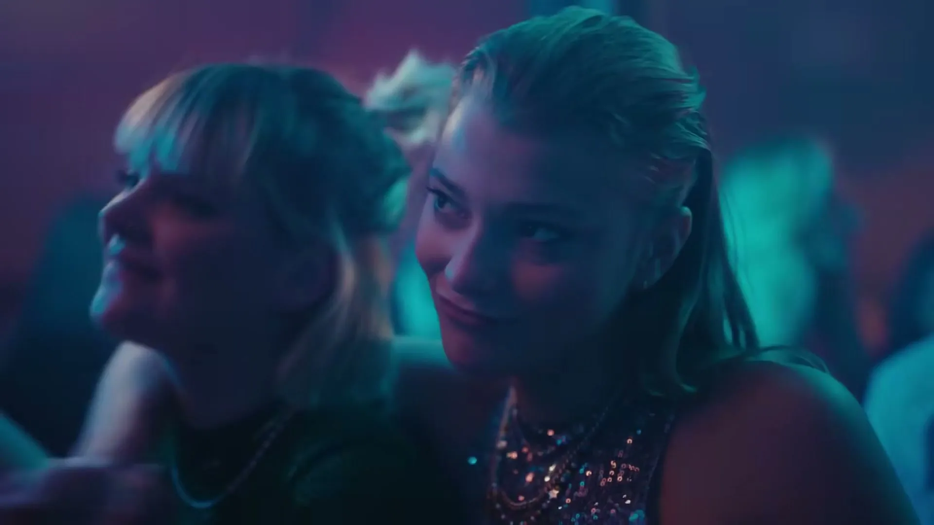 Two young women, one with blonde hair and the other with brown hair, dance closely together in a dimly lit club, smiling and embracing.