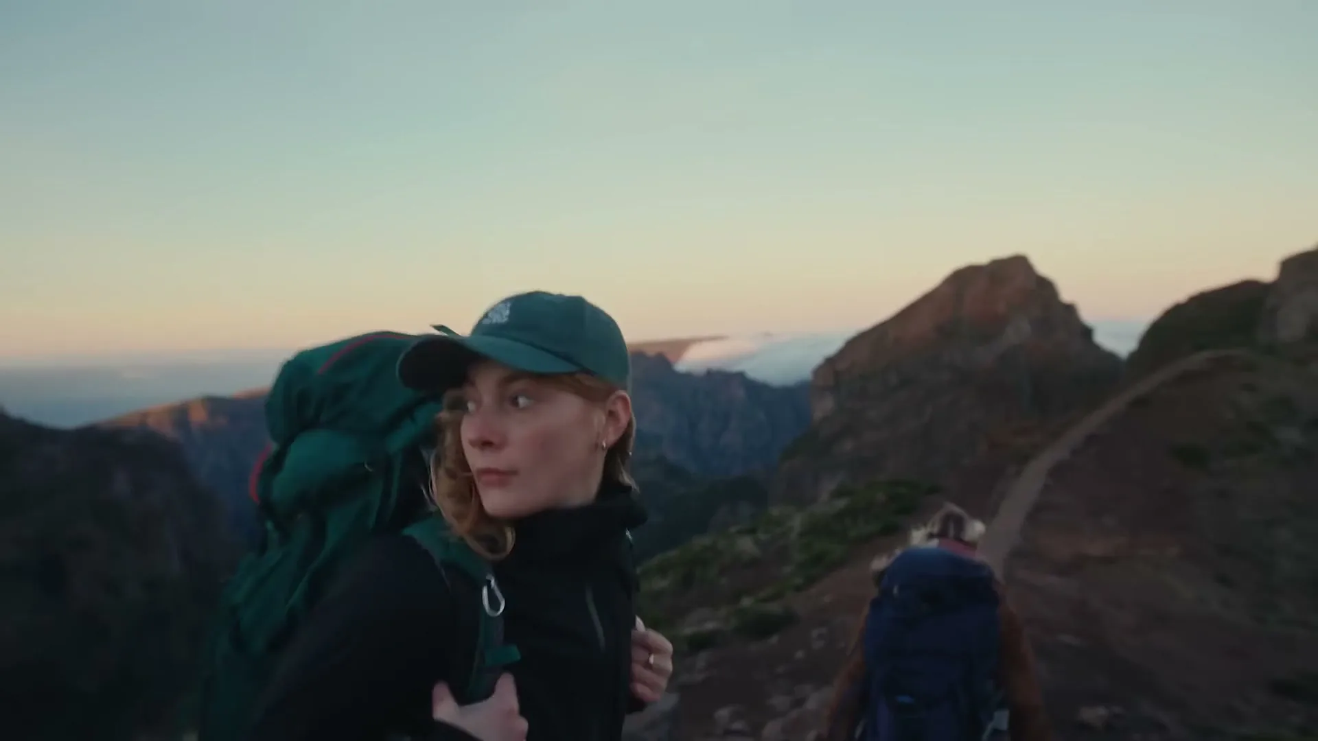A young woman with a green backpack and cap hikes on a mountain trail at dawn, looking back at another hiker.
