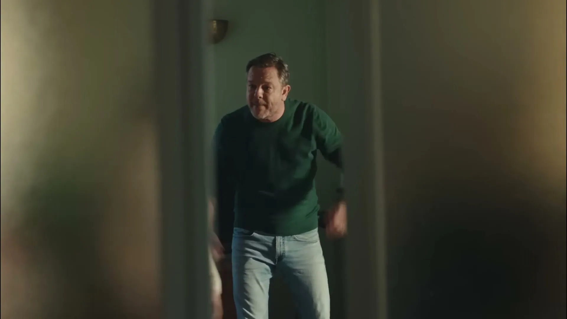 A man in a green sweater and jeans shouts at a young girl who covers her ears, seen through a doorway.