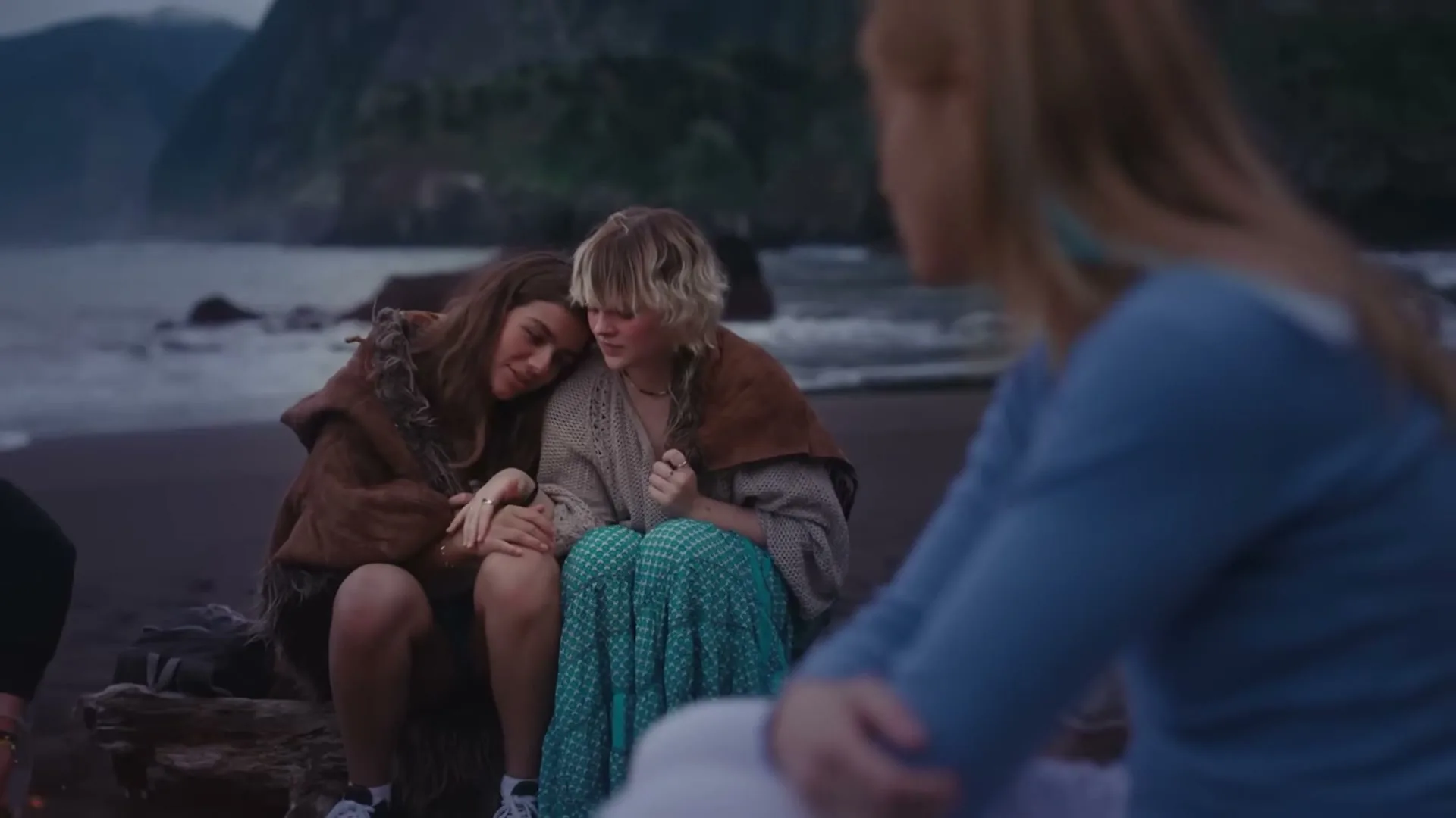 Two young women huddle together for warmth by a campfire on a dark beach, while another woman sits in the foreground.
