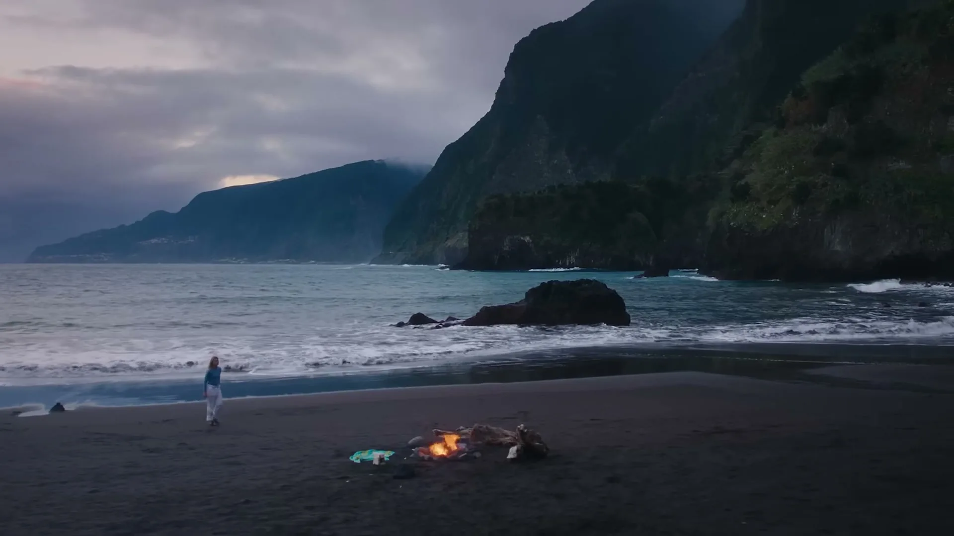 A woman walks along a dark sand beach with a small bonfire, as waves crash against the shore and large, dark mountains rise in the background under a cloudy sky.