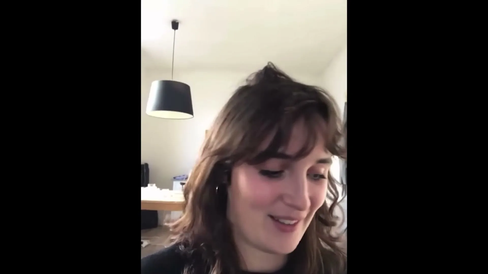 A young woman with brown hair and a black top smiles and looks down, possibly at a phone, in a brightly lit room.