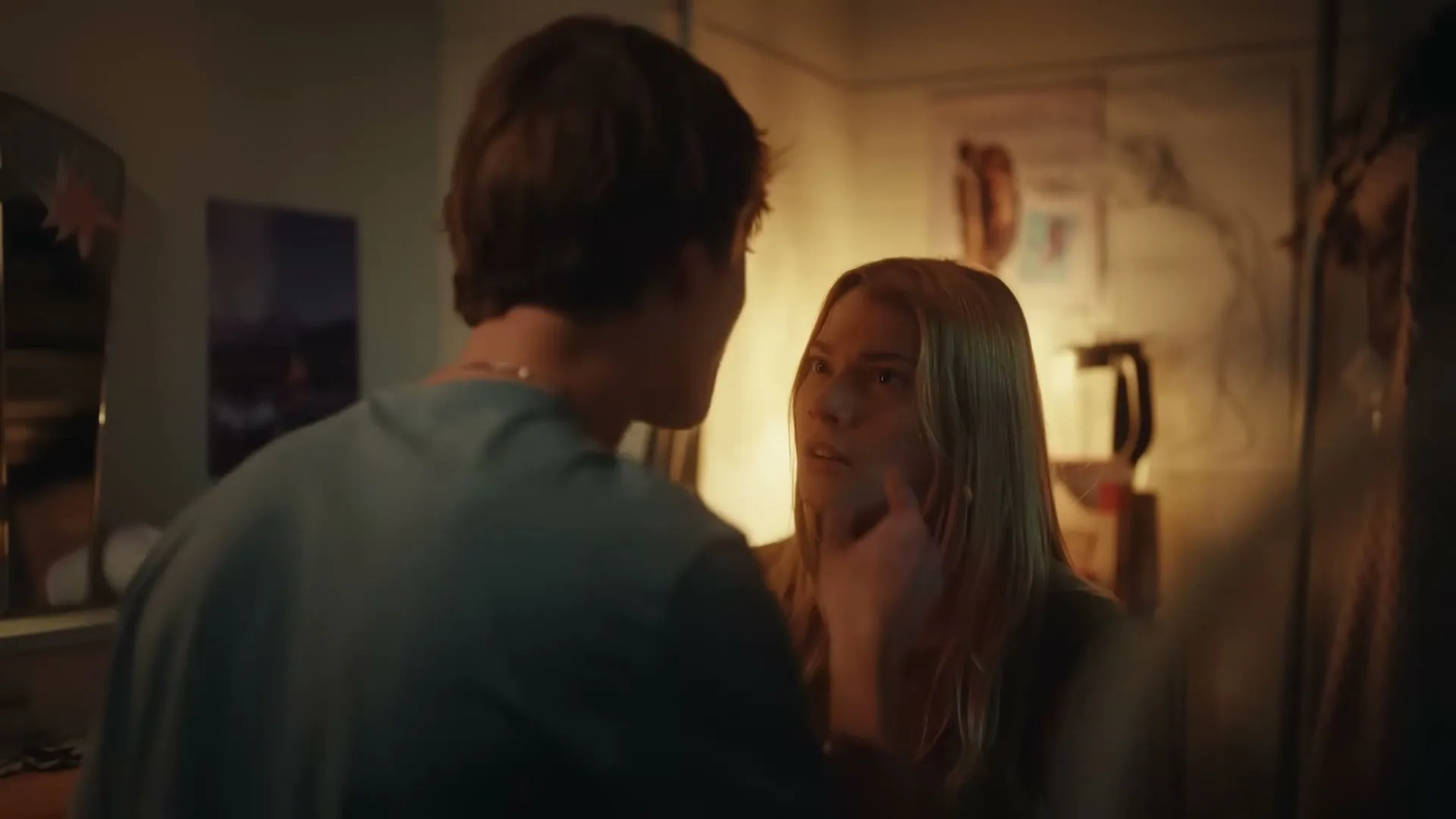 A young man and woman stand close, looking at each other with concern in a dimly lit room.
