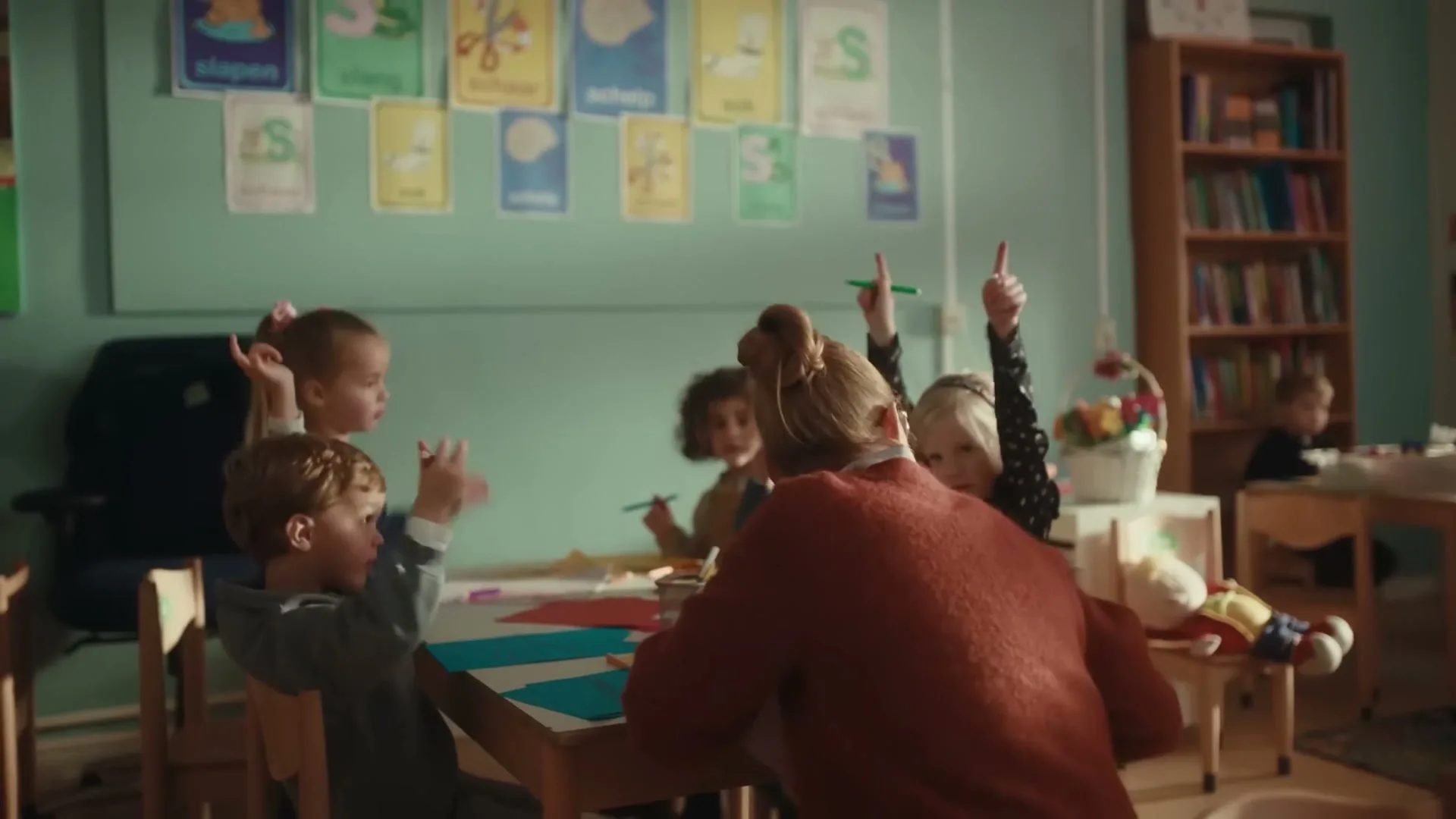 A female teacher with a bun sits at a table with four young children, who raise their hands in a brightly lit classroom.