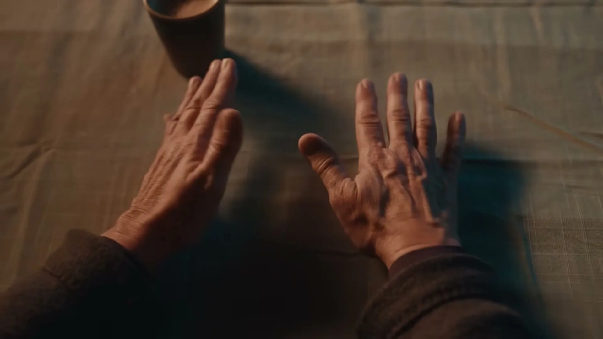 An older person's hands are shown palms up on a table, then slowly turn palms down, next to a cup.