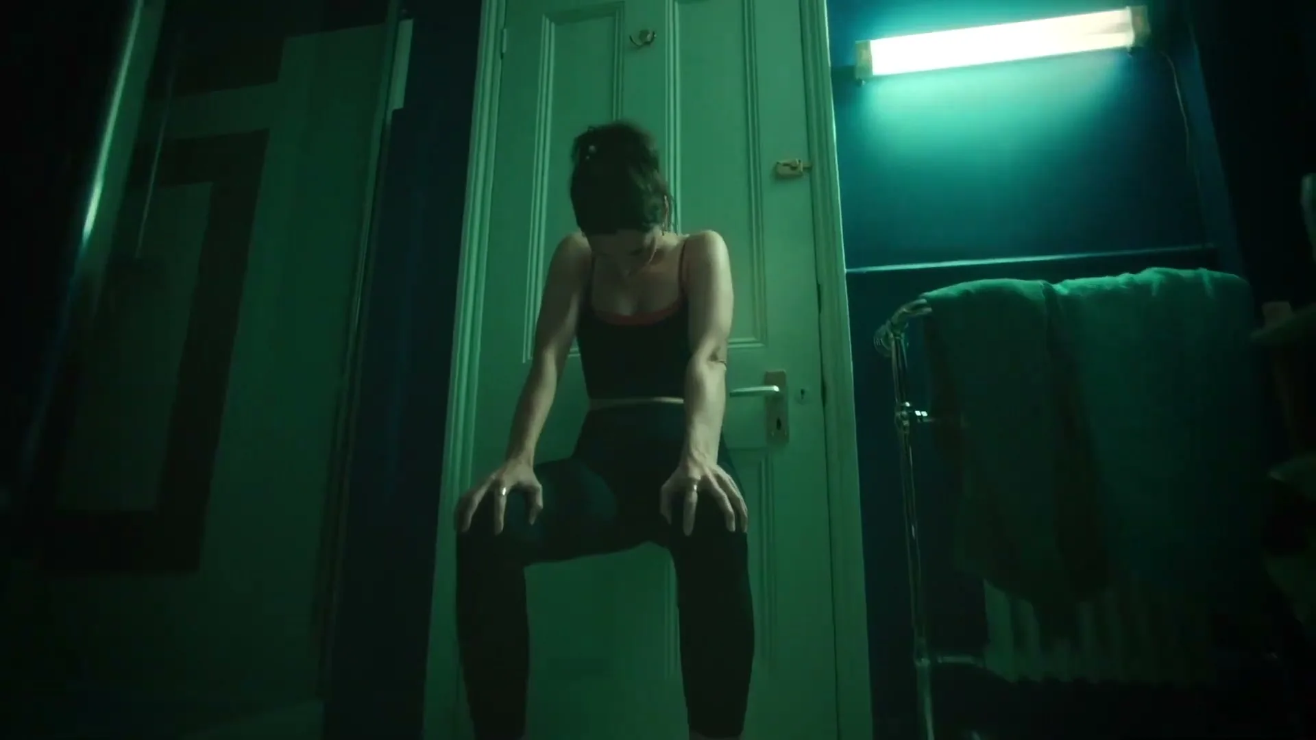 A woman in workout clothes sits on the floor, leaning against a door in a dimly lit, teal-colored bathroom, looking exhausted.