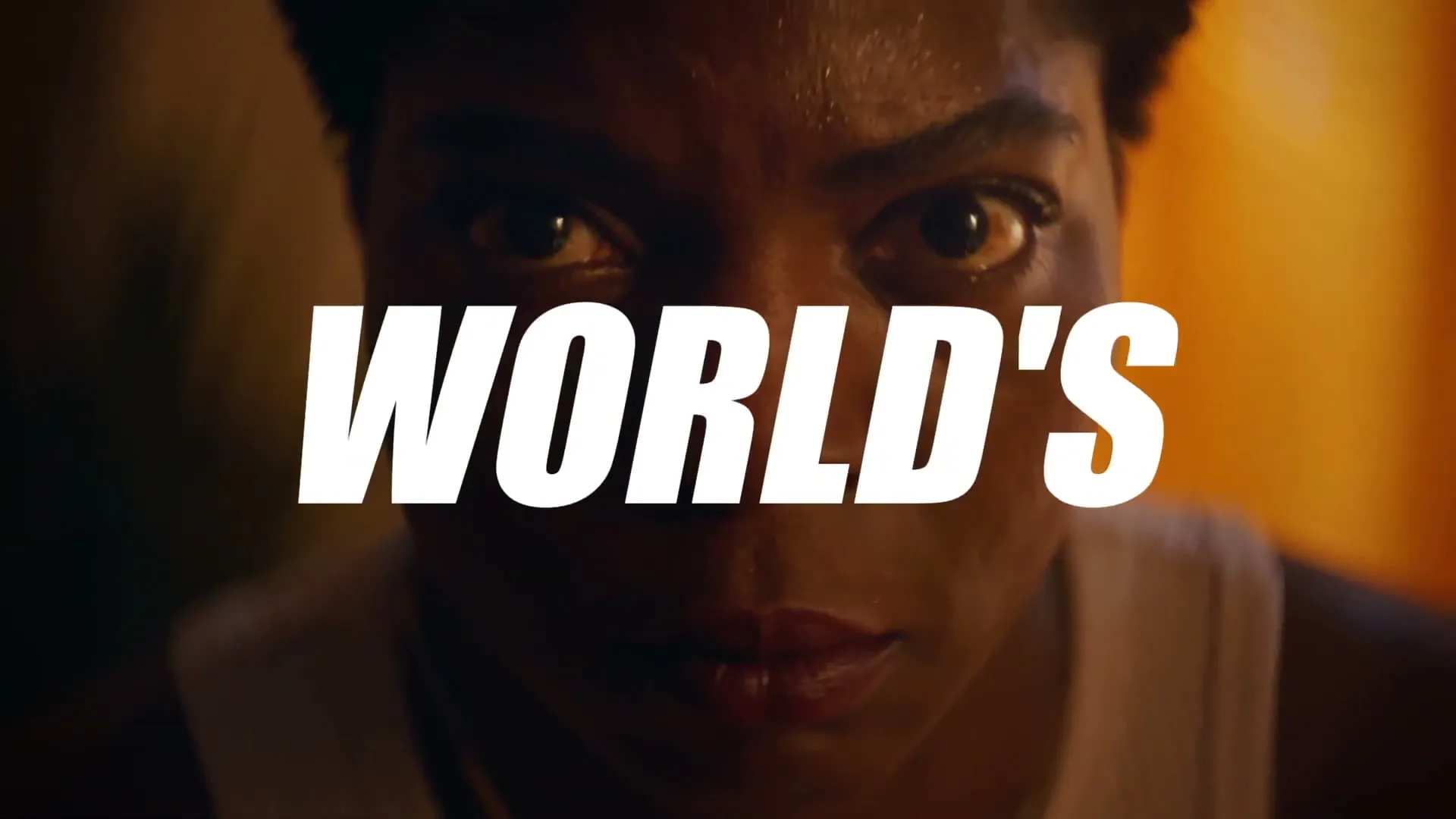 A determined Black woman in her 30s, wearing a white tank top, looks intensely at the camera with sweat on her face, as the word "WORLD'S" appears over her eyes.