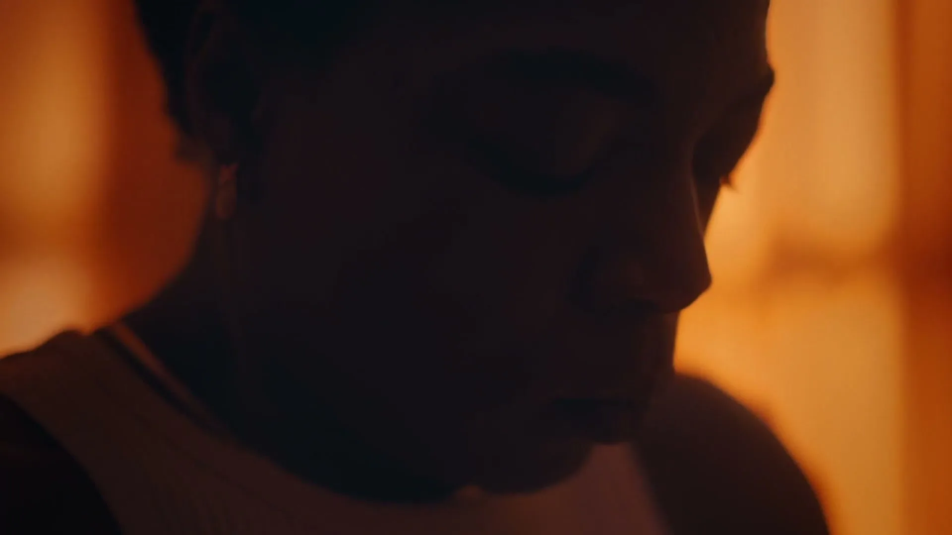 A young woman with short hair is seen in a dark silhouette against a warm, blurry background, looking down thoughtfully.