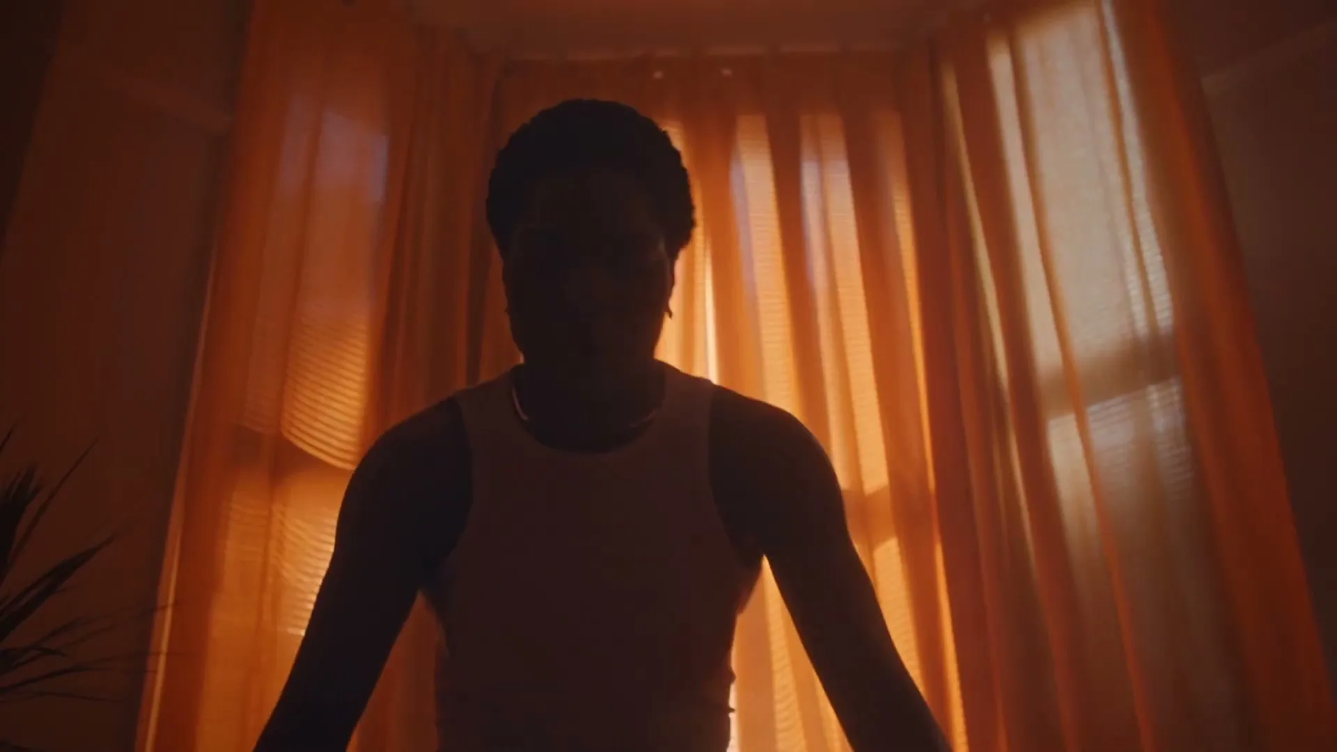 A young Black woman in a tank top is silhouetted against orange curtains, looking directly at the camera in an interior setting.