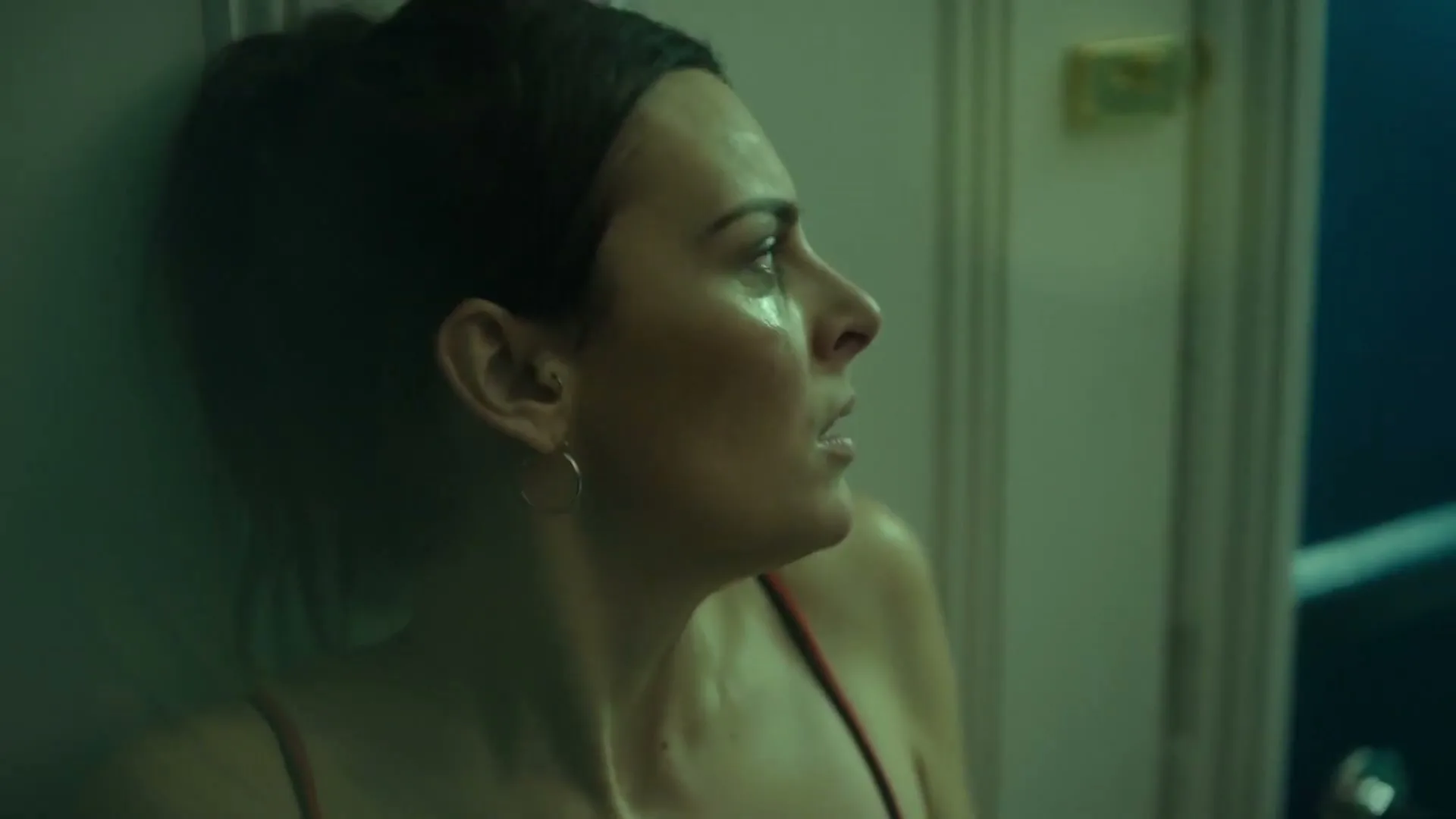 A woman with dark hair, wearing a red top, leans against a wall, looking distressed in a dimly lit room.