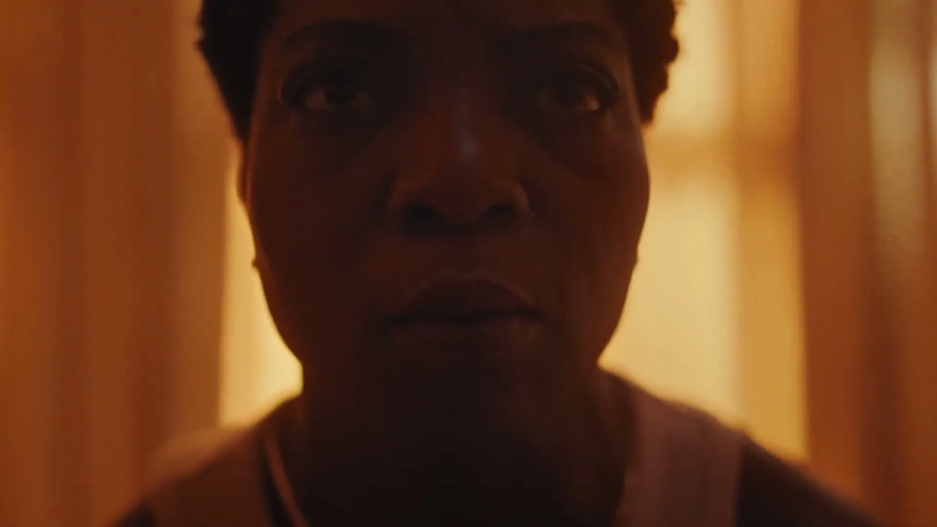 A woman with short dark hair, wearing a white tank top, slowly opens her eyes and looks up, in a dimly lit room.