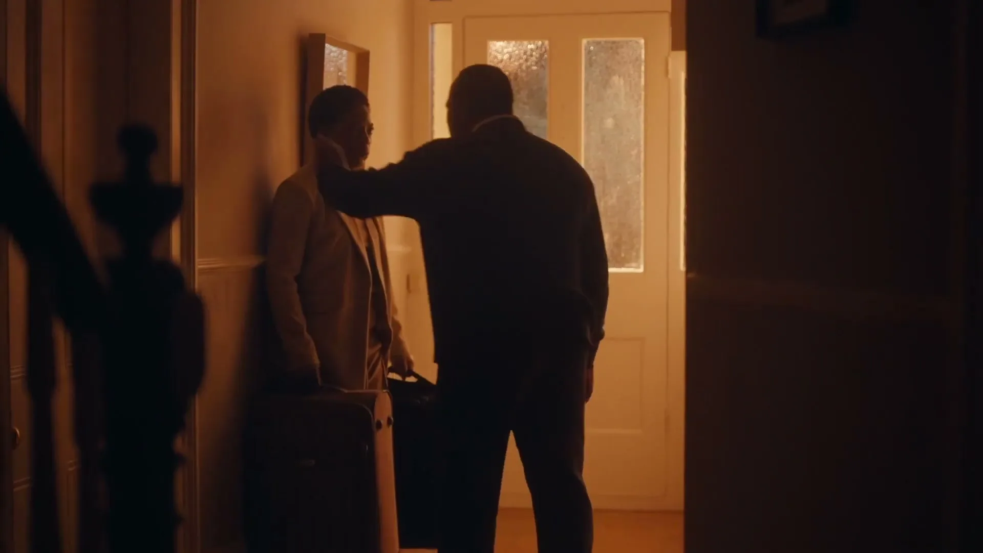 A man and a woman stand in a dimly lit hallway, the woman holding a suitcase, as the man gently touches her face before she leaves.