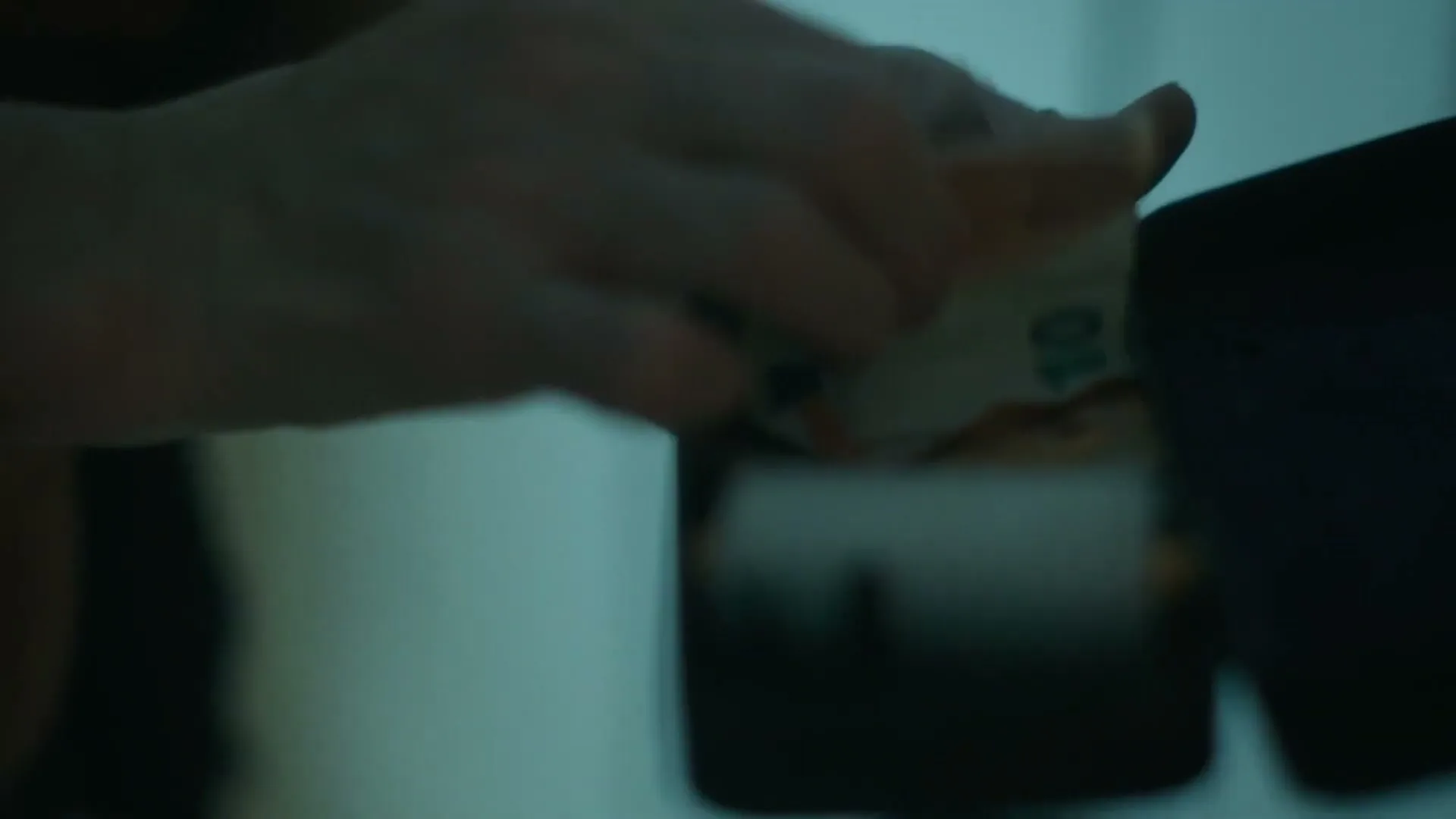 A person's hands are seen in a medium closeup, counting and placing euro banknotes into a dark wallet in a dimly lit interior.