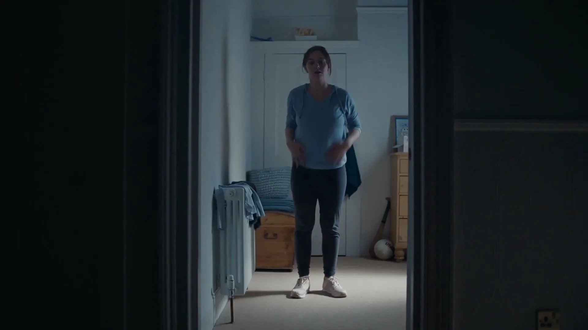A woman in her 30s stands in a dimly lit bedroom doorway, looking surprised and then adjusting her clothes.