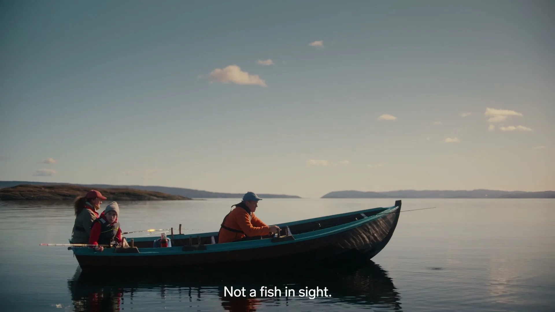 A man, woman, and child are fishing from a small boat on a calm lake with distant land under a clear sky.