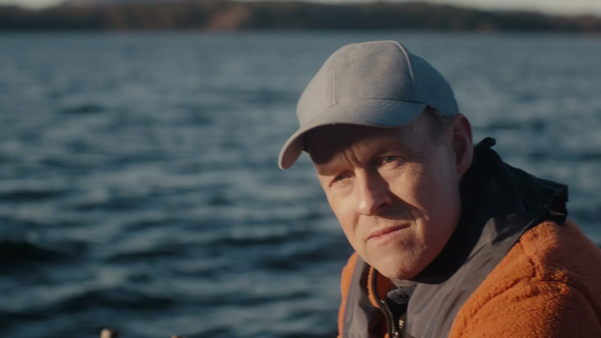 A man in a grey cap and orange fleece jacket looks out over the water, then turns to face the camera on a boat during the day.