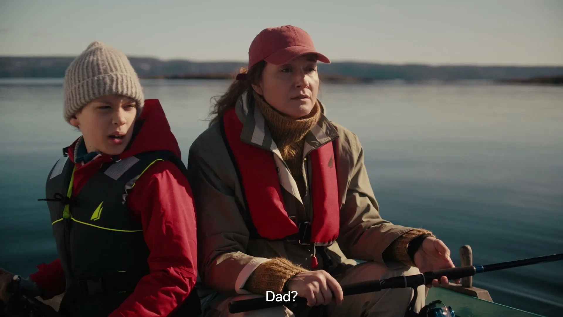 A young boy and a woman in a red cap and life vests sit in a small boat on a calm lake, looking off-camera with fishing rods in hand.