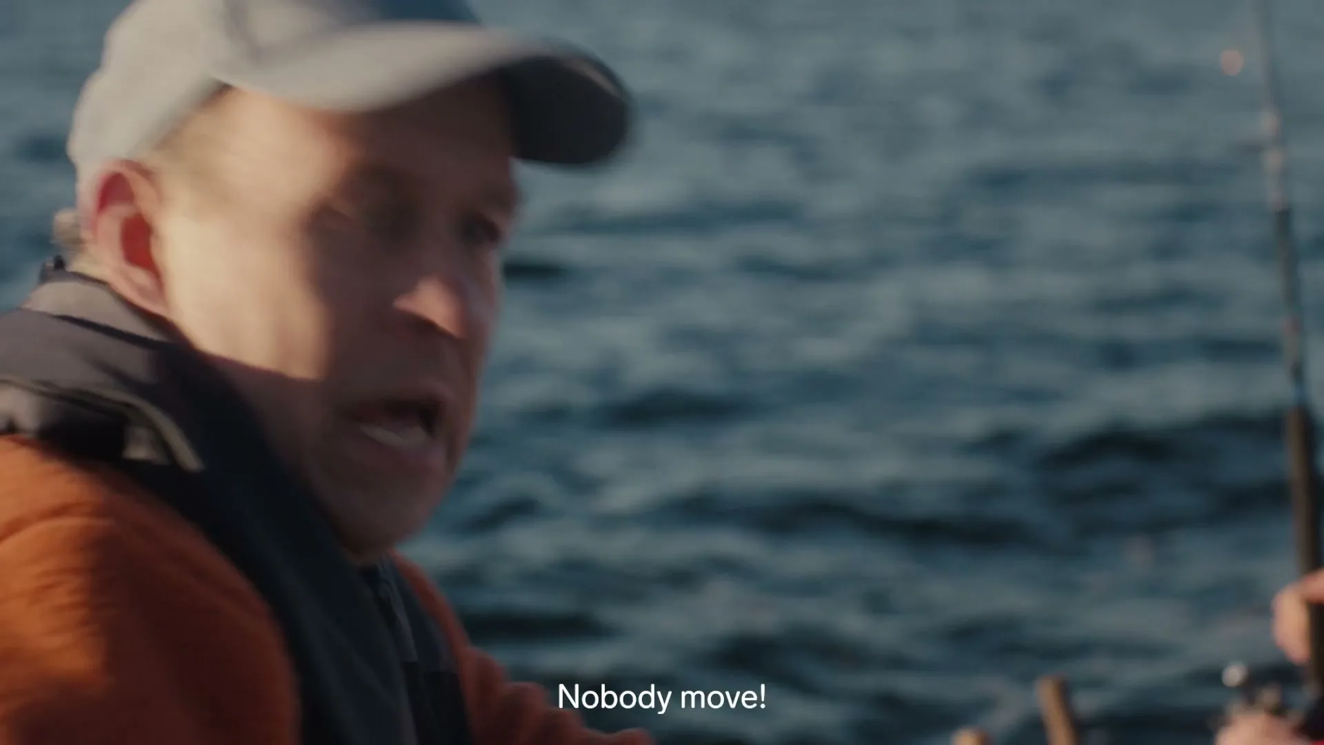 A man in a grey cap and orange jacket looks over his shoulder at something in the water, then turns to shout at someone off-screen on a boat.