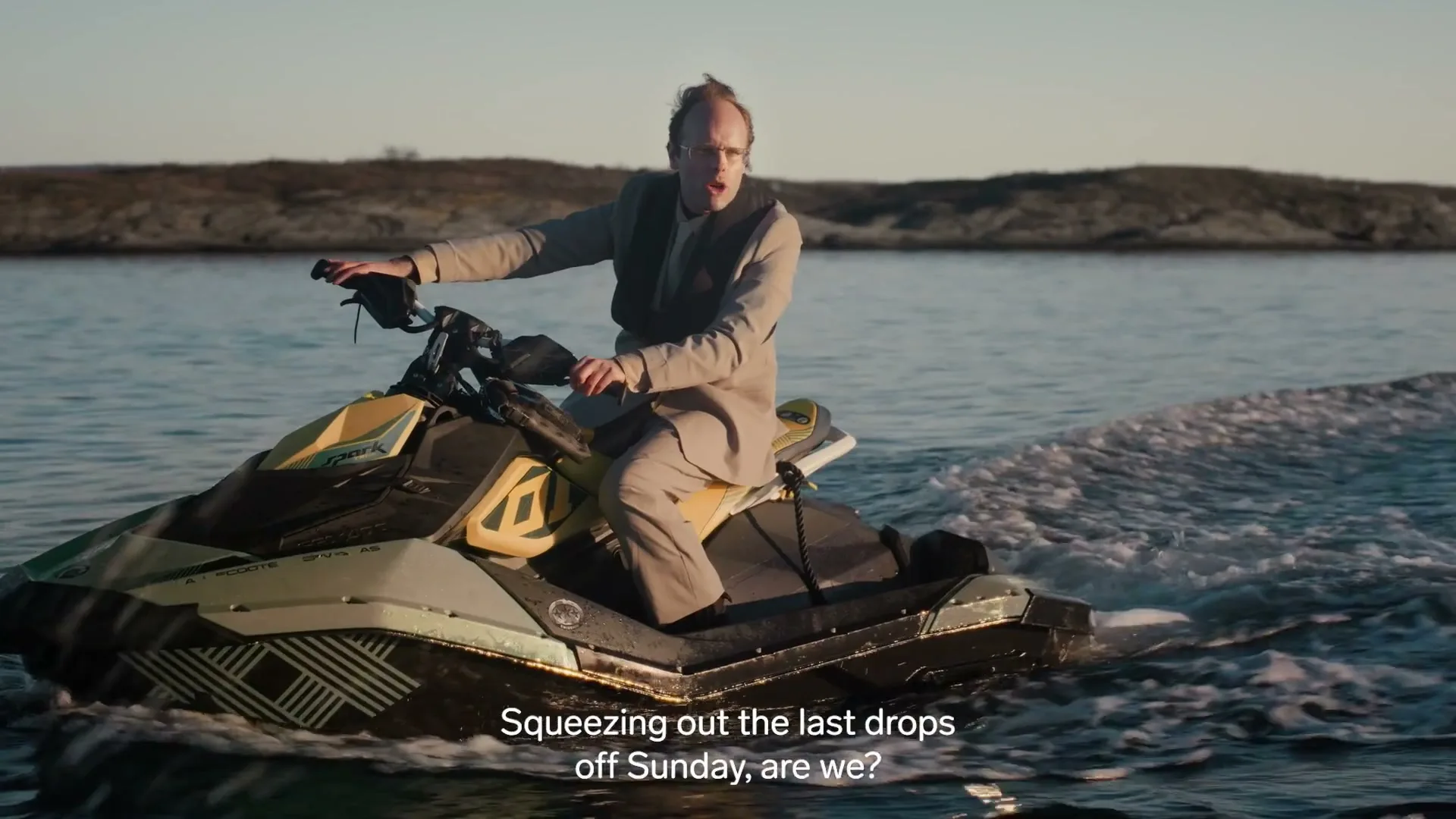 A man in a suit and life vest rides a jet ski on the water, with a rocky coastline in the background under a clear sky.