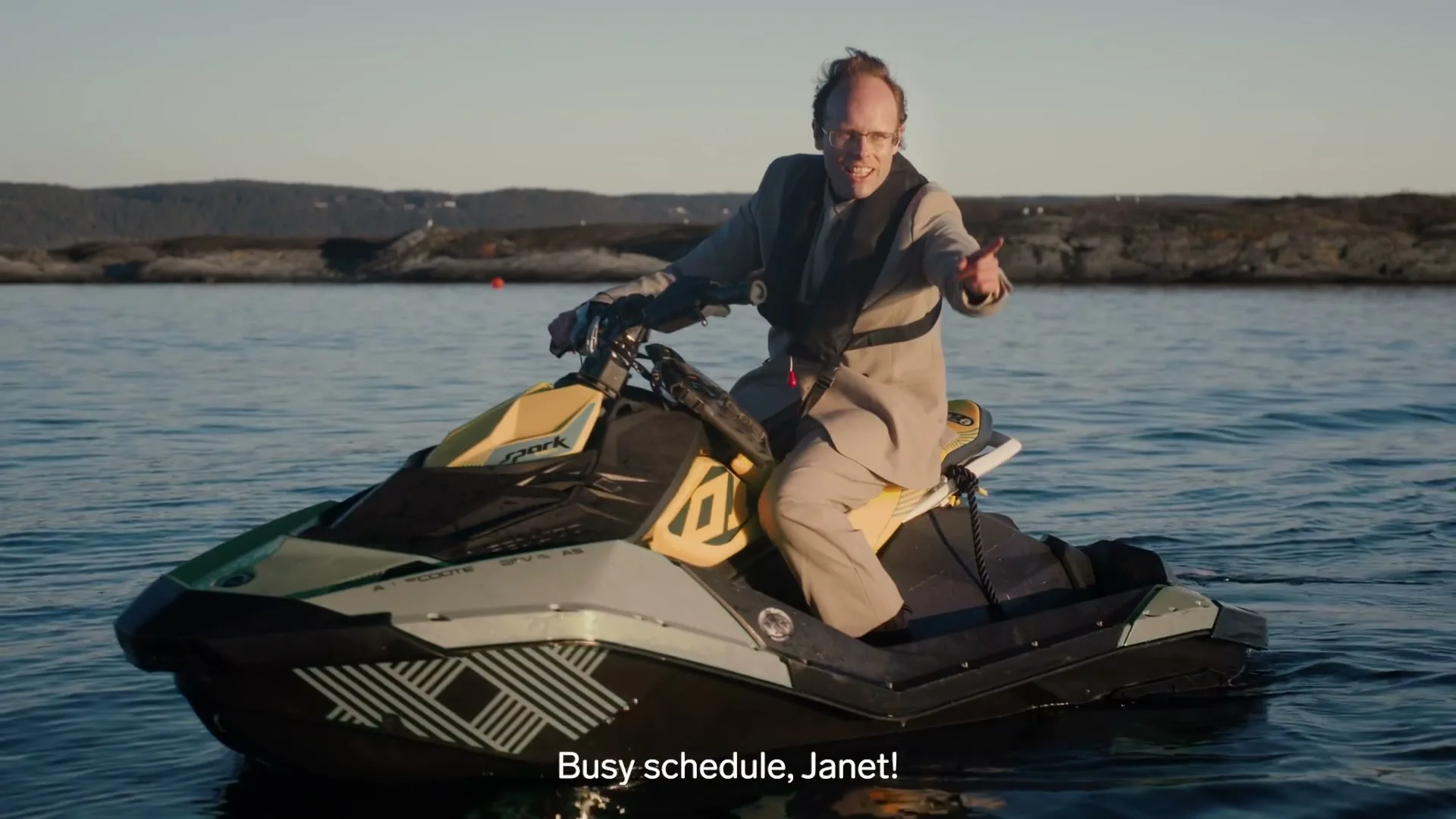 A man in a suit and life vest rides a jet ski on calm water, pointing and smiling at the camera with a rocky coastline in the background.