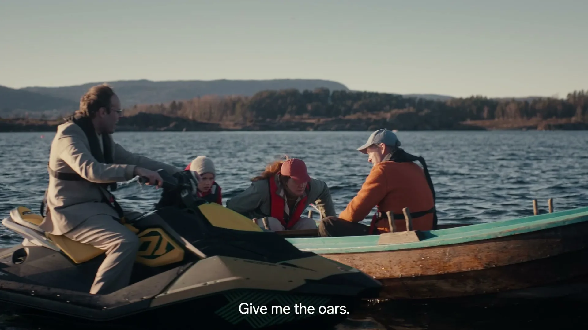 A man in a suit on a jet ski talks to a family in a small boat on a lake, with a forested shore in the background.