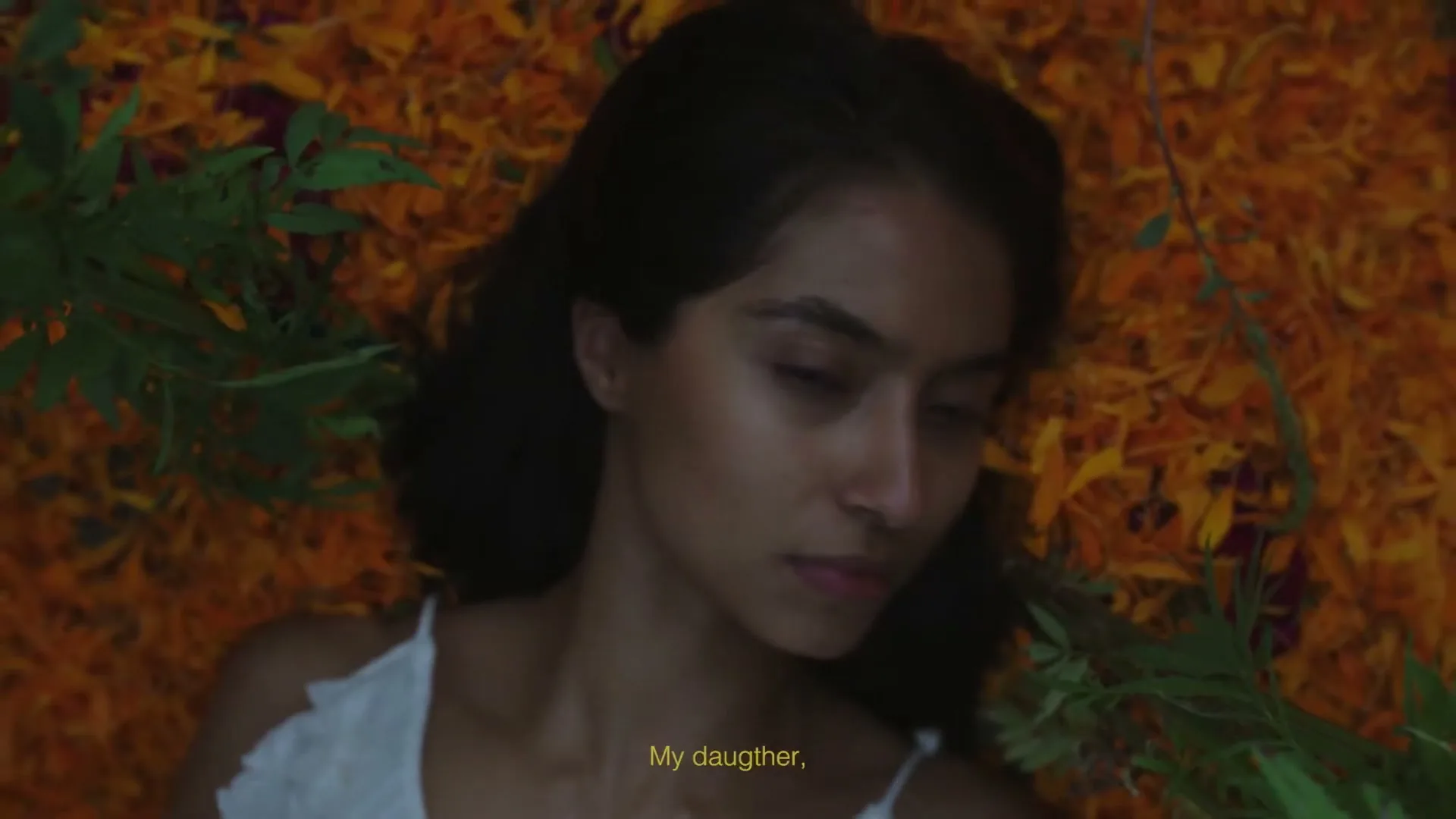 A young woman with dark hair and a white top lies on a bed of orange marigold petals and green leaves, opening her eyes slowly.
