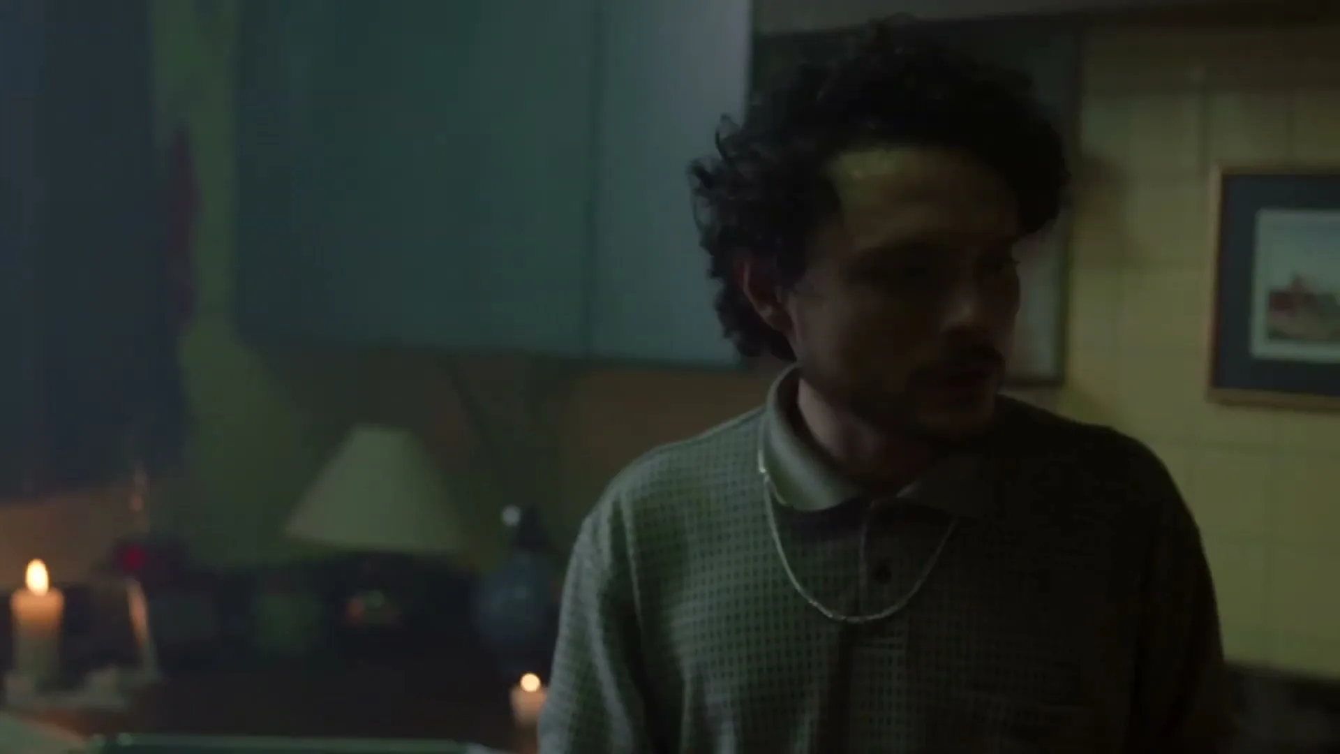 A young man with curly hair and a polo shirt looks around a dimly lit room with candles and a lamp.