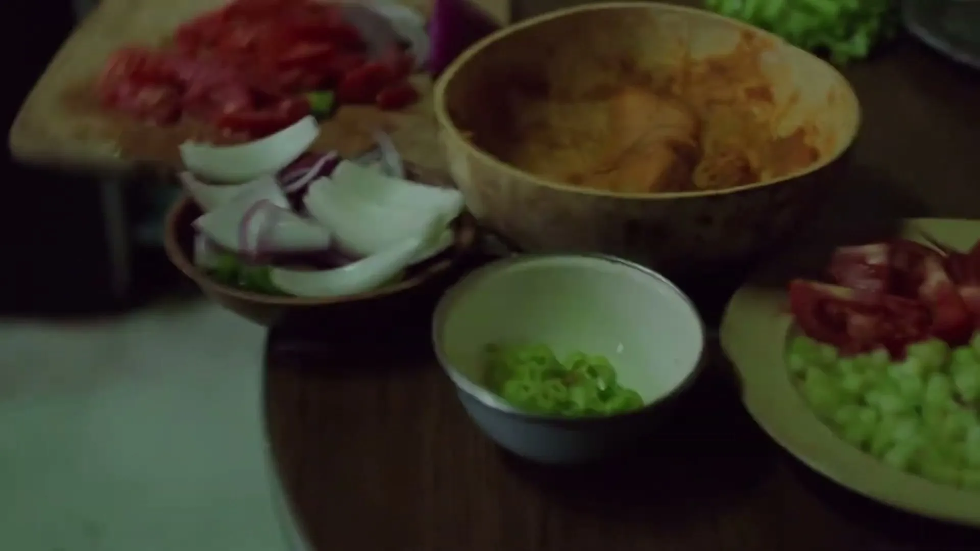 A close-up shot shows various bowls and plates filled with chopped vegetables, including tomatoes, onions, and green peppers, on a wooden table in a kitchen.