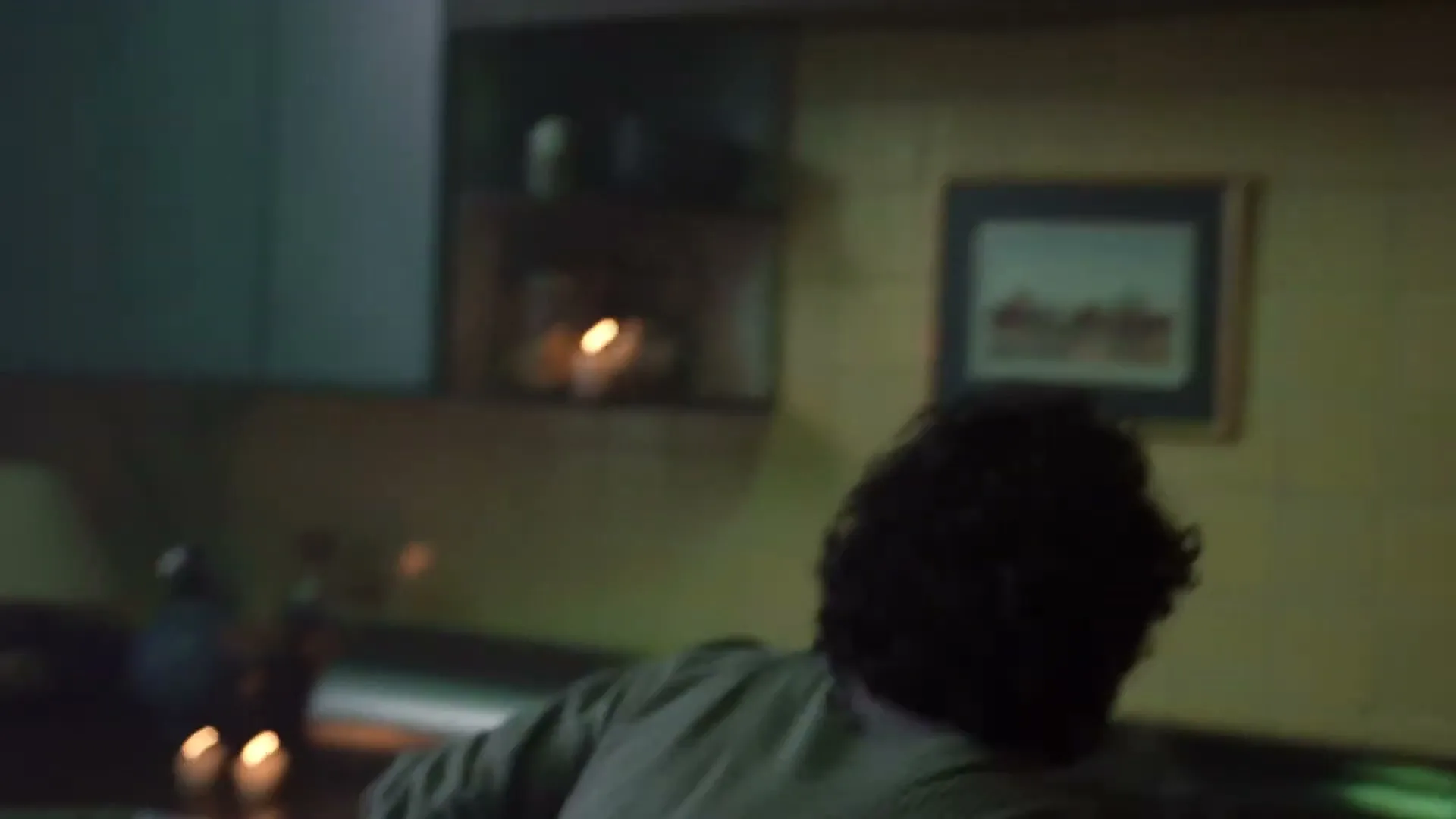 A man with dark, curly hair and a striped shirt leans over a counter in a dimly lit kitchen, looking down intently.