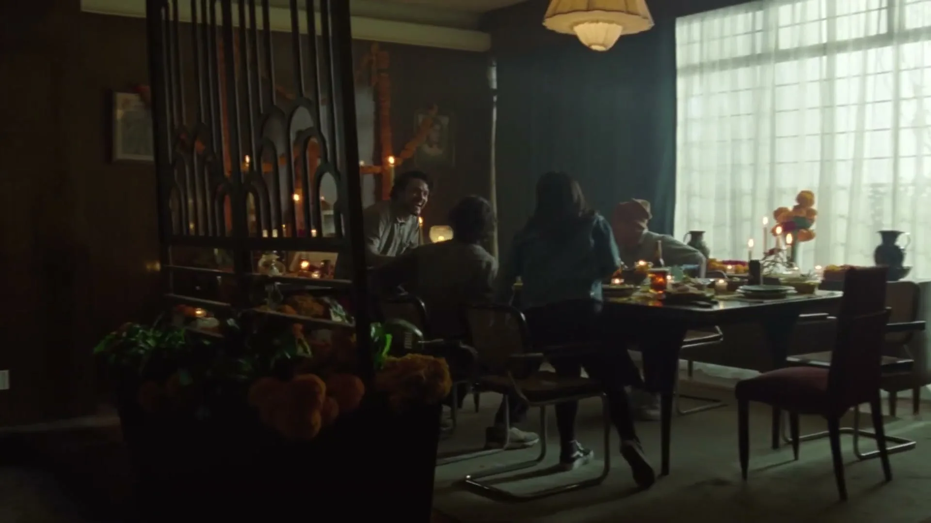 A group of people gather around a dining table, decorated with candles and flowers, in a dimly lit room with a large window.