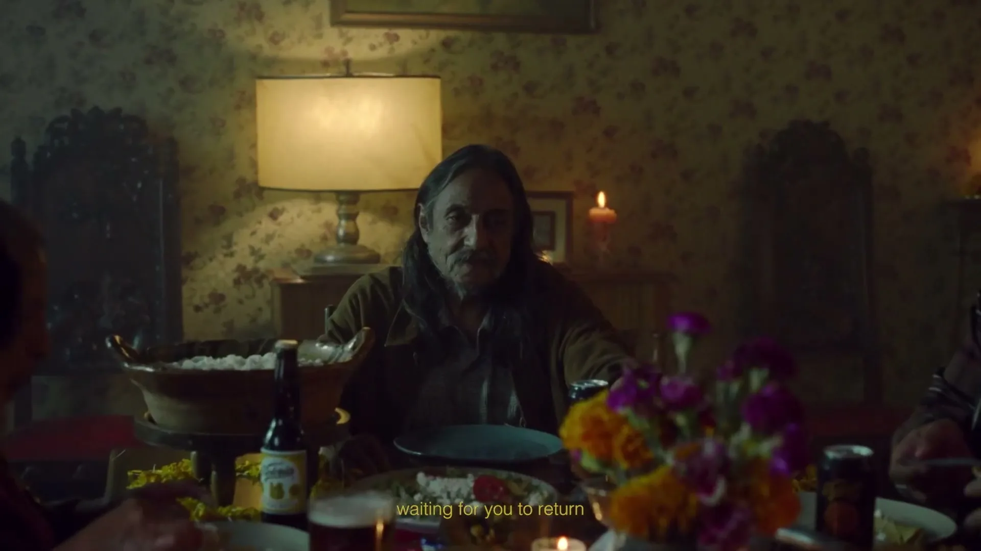 A man with long hair and a mustache sits at a dimly lit dinner table, looking forward with a neutral expression.