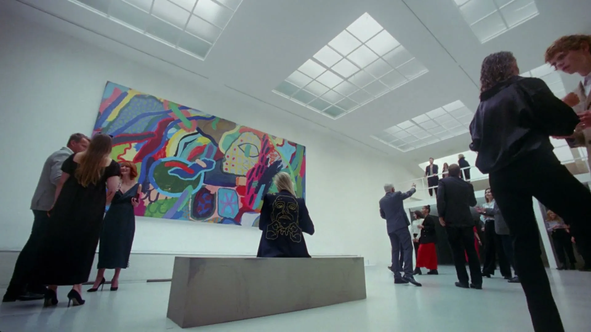 A diverse group of people in formal attire mingle and observe a large, colorful abstract painting in a modern art gallery.