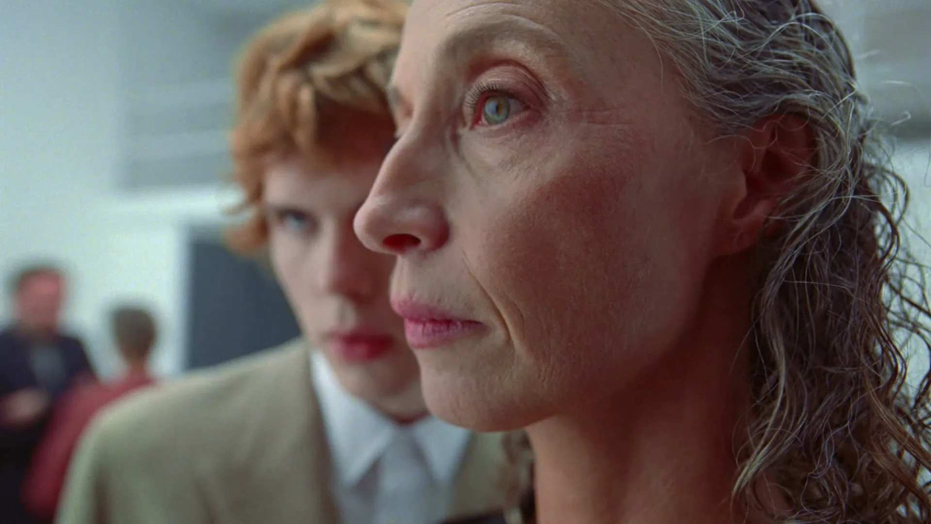 A woman with gray hair and a young man with red hair stand in an interior space, with the woman looking to her right.