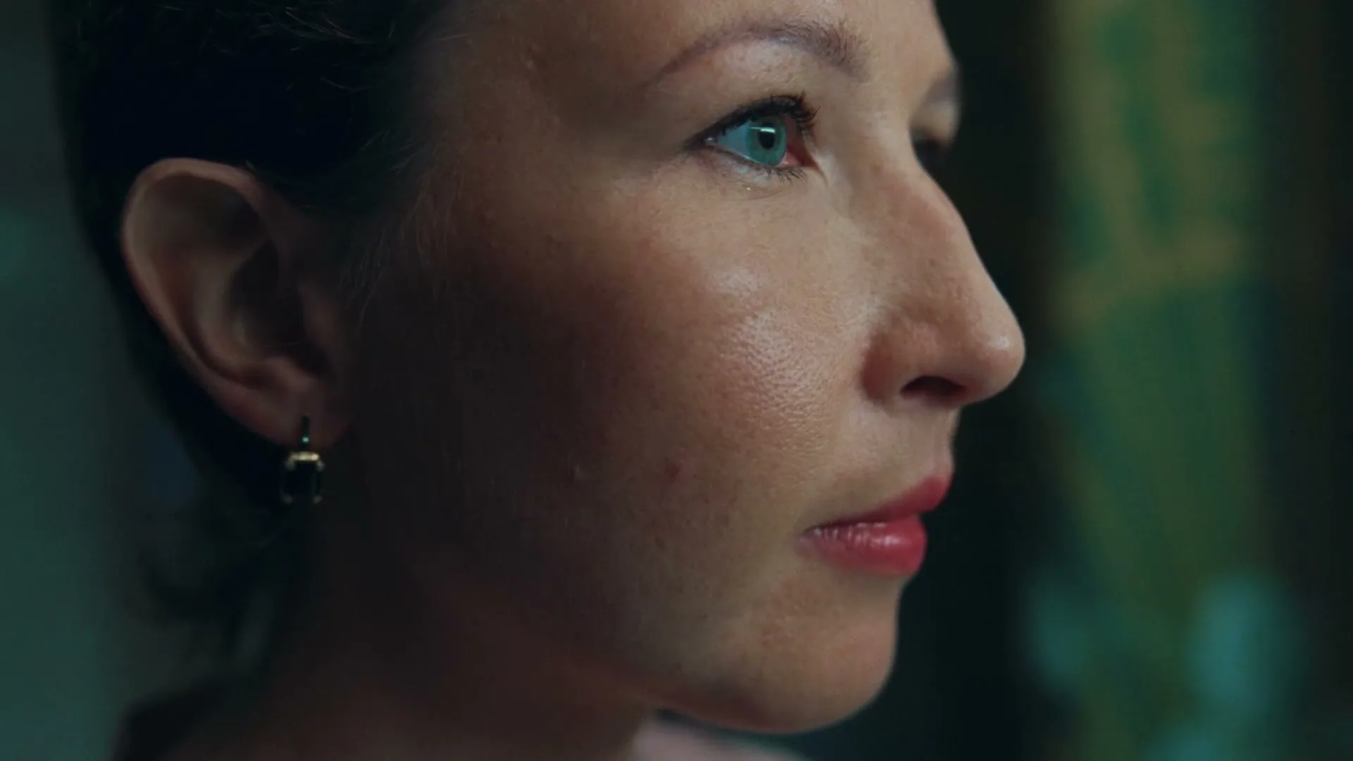 A woman with blue eyes and red lipstick looks to the right, then puts on an earring.