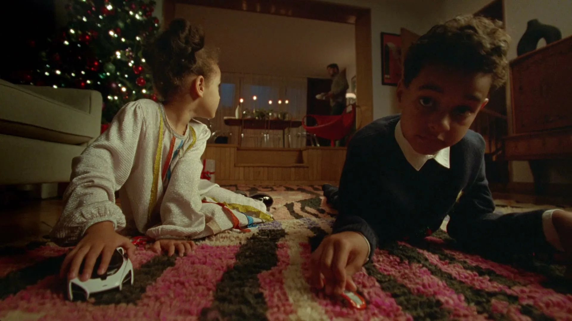Two children, a girl and a boy, play with toy cars on a patterned rug in a living room with a Christmas tree.