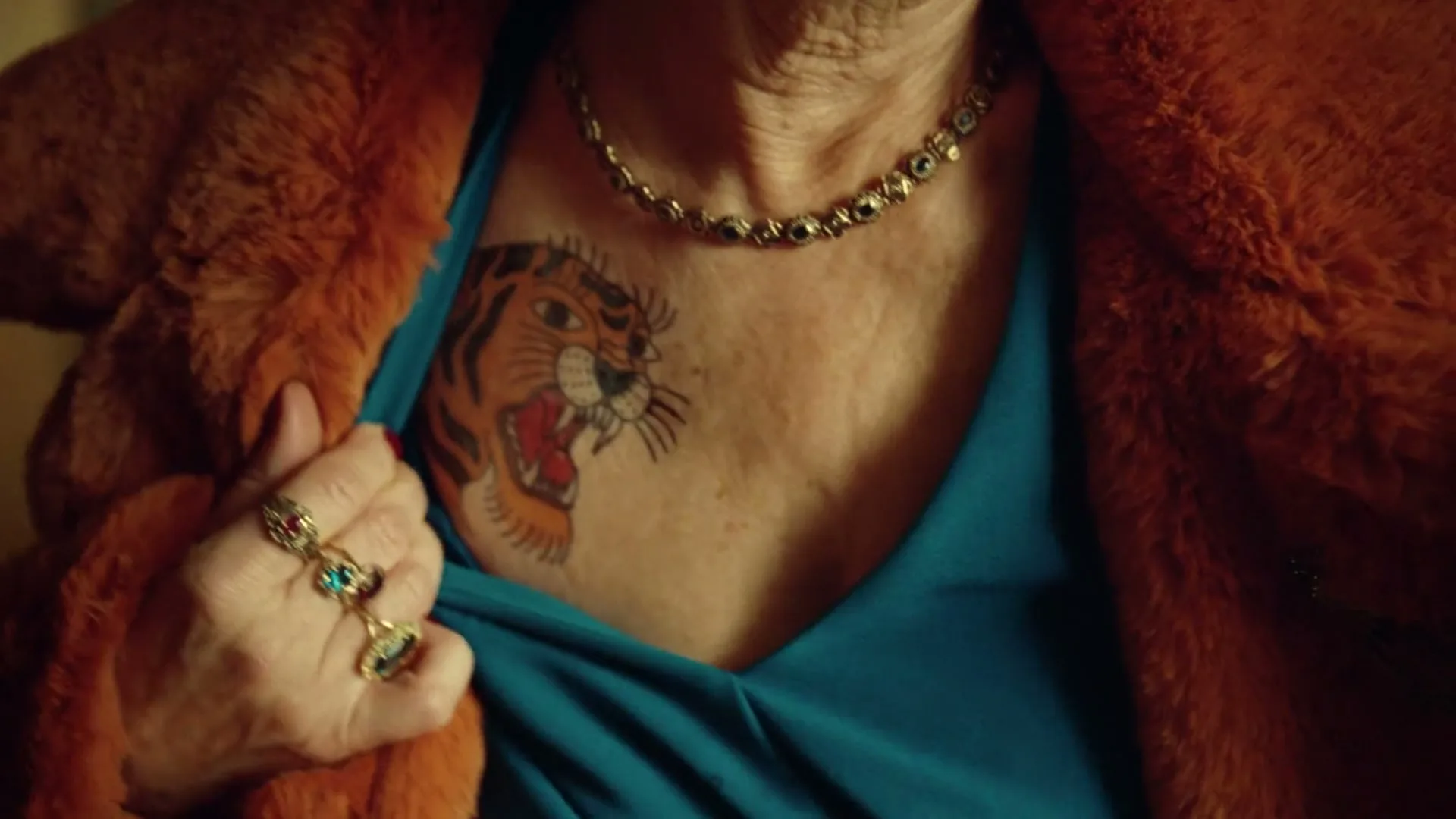 A close-up shot of an older woman's chest, revealing a tiger tattoo and a blue top under an orange fur coat.