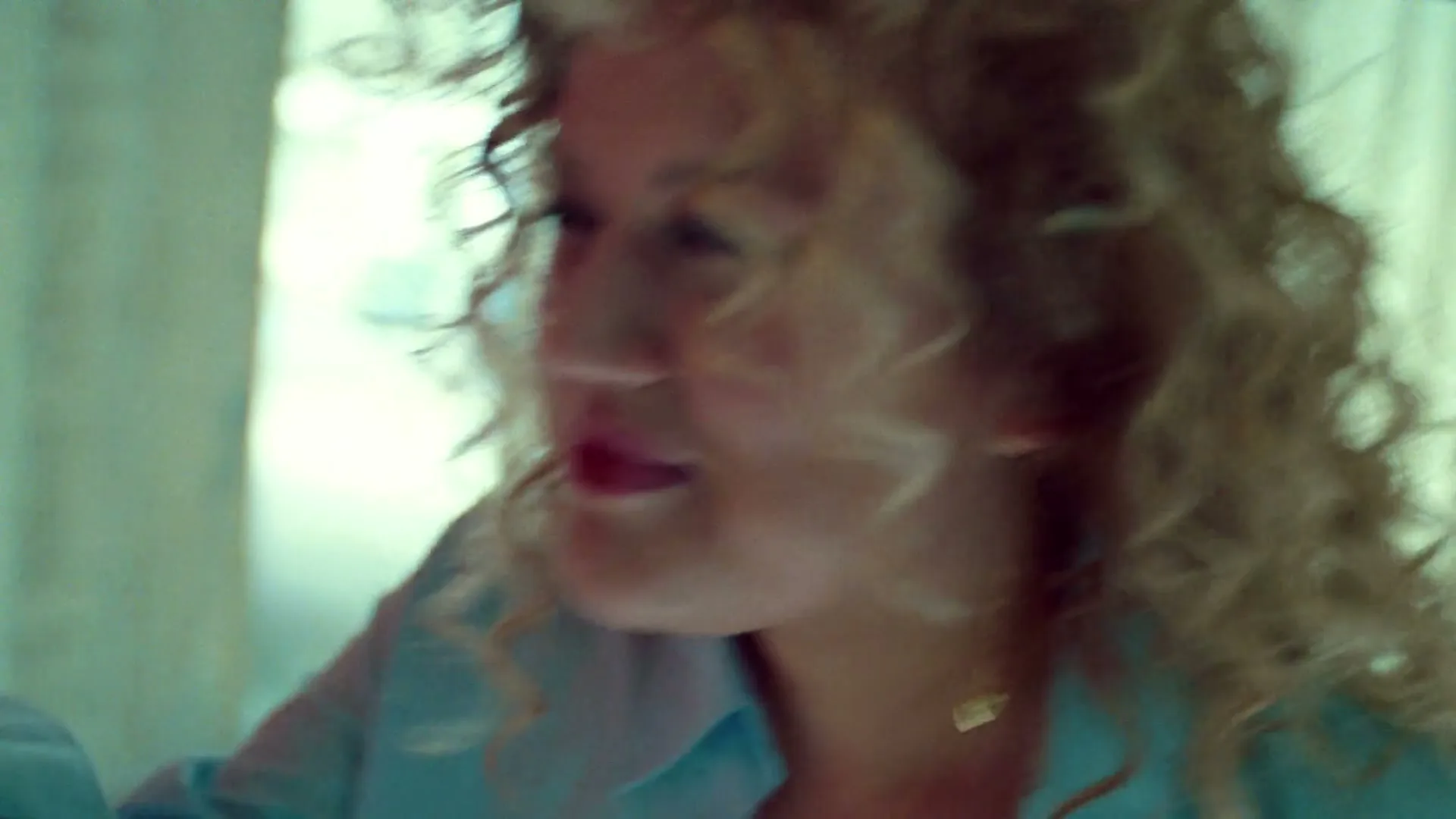 A woman with curly blonde hair and red lipstick looks to the side, then reaches for a red door handle in an interior setting.