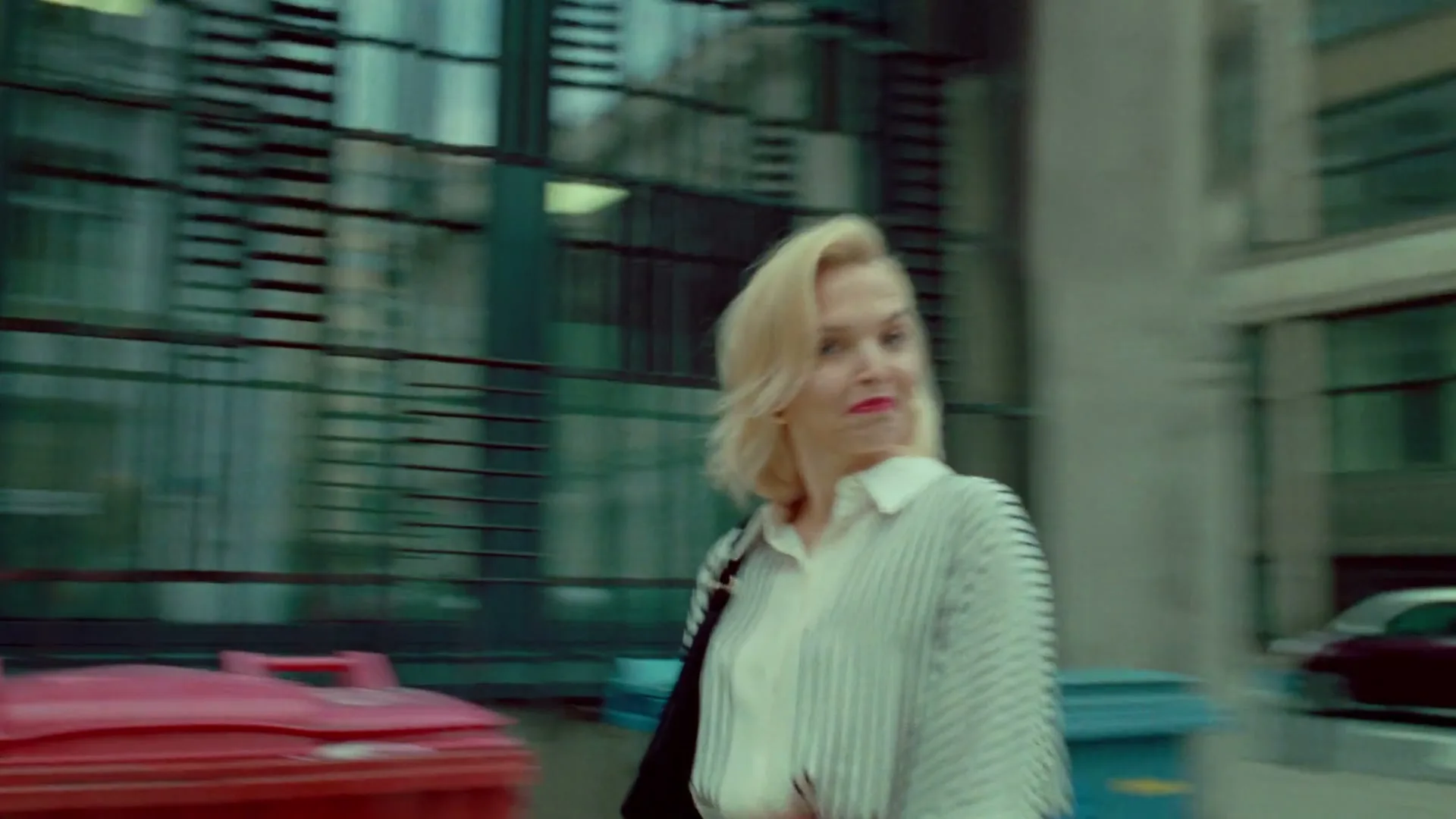 A blonde woman in a striped shirt and red skirt walks past a blue dumpster on a city street during the day.