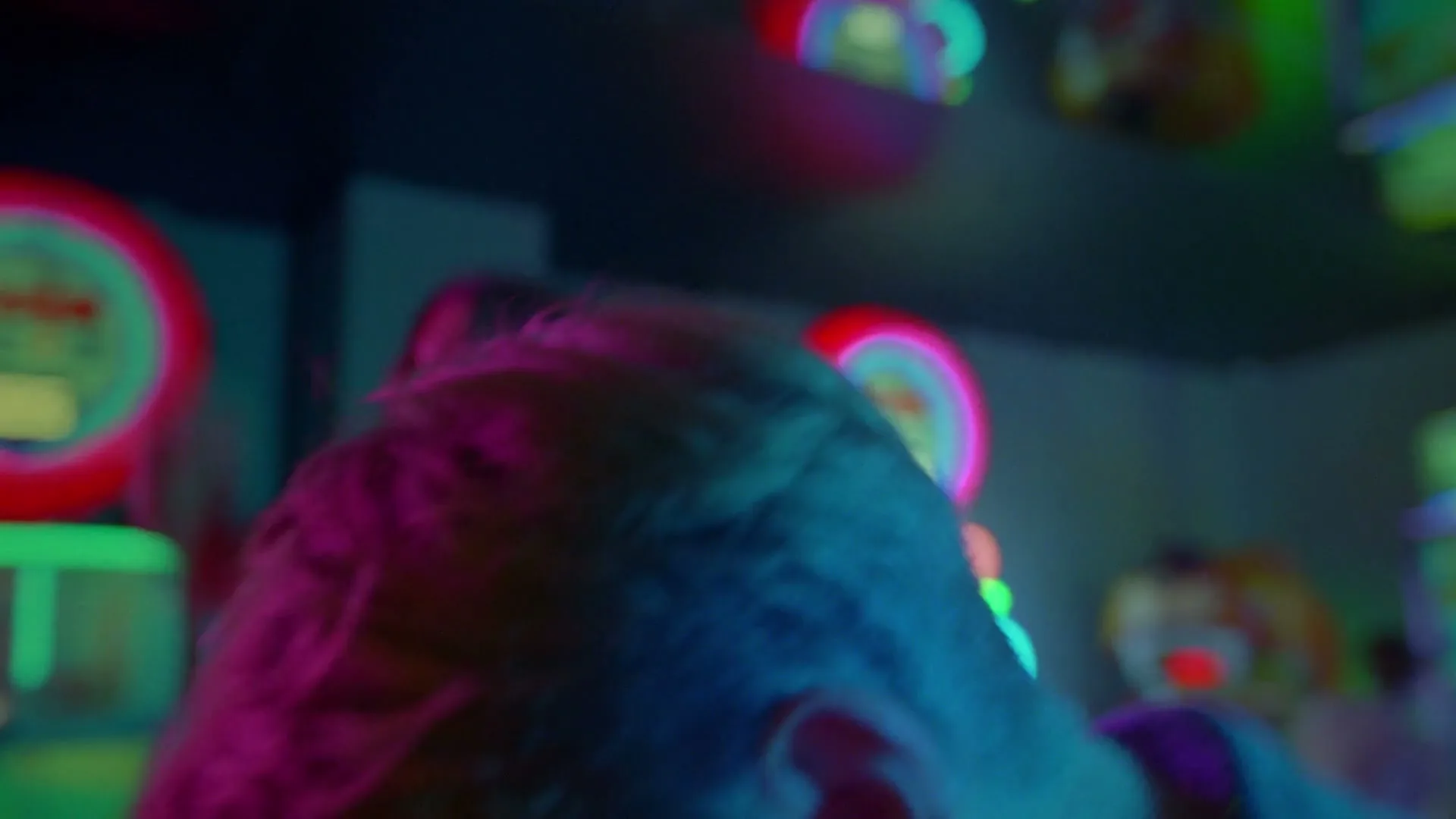 A young woman with short blonde hair, wearing a denim jacket, laughs and dances in a neon-lit arcade.