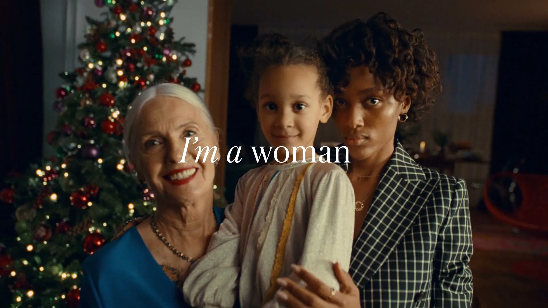 Three females of different generations stand together in front of a Christmas tree, looking at the camera.