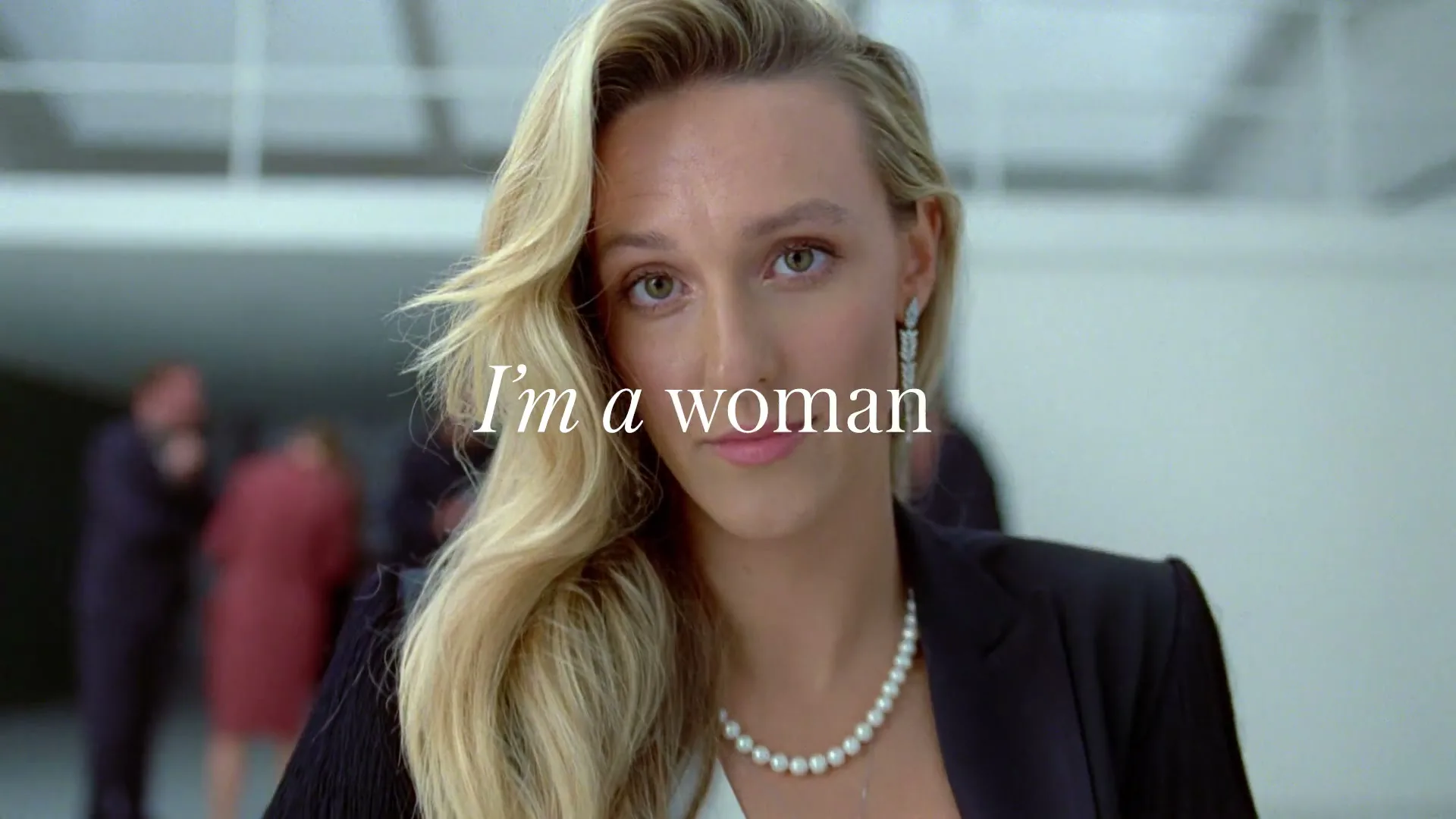 A blonde woman in a black blazer and pearl necklace looks directly at the camera with a slight smile, with "I'm a woman" text overlay.