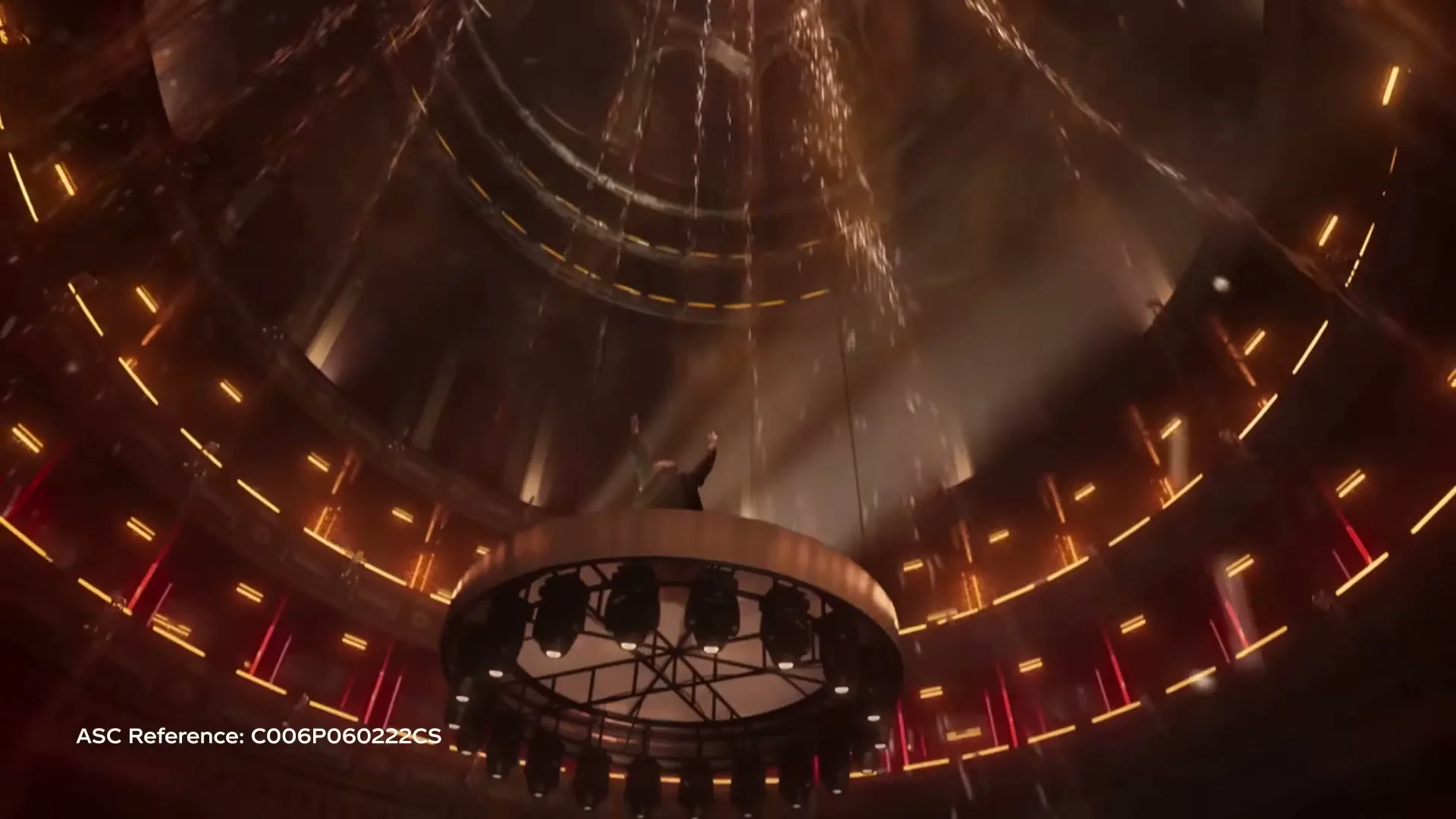 A man in a dark suit conducts an orchestra in a grand, circular concert hall, with golden lights and falling glitter.